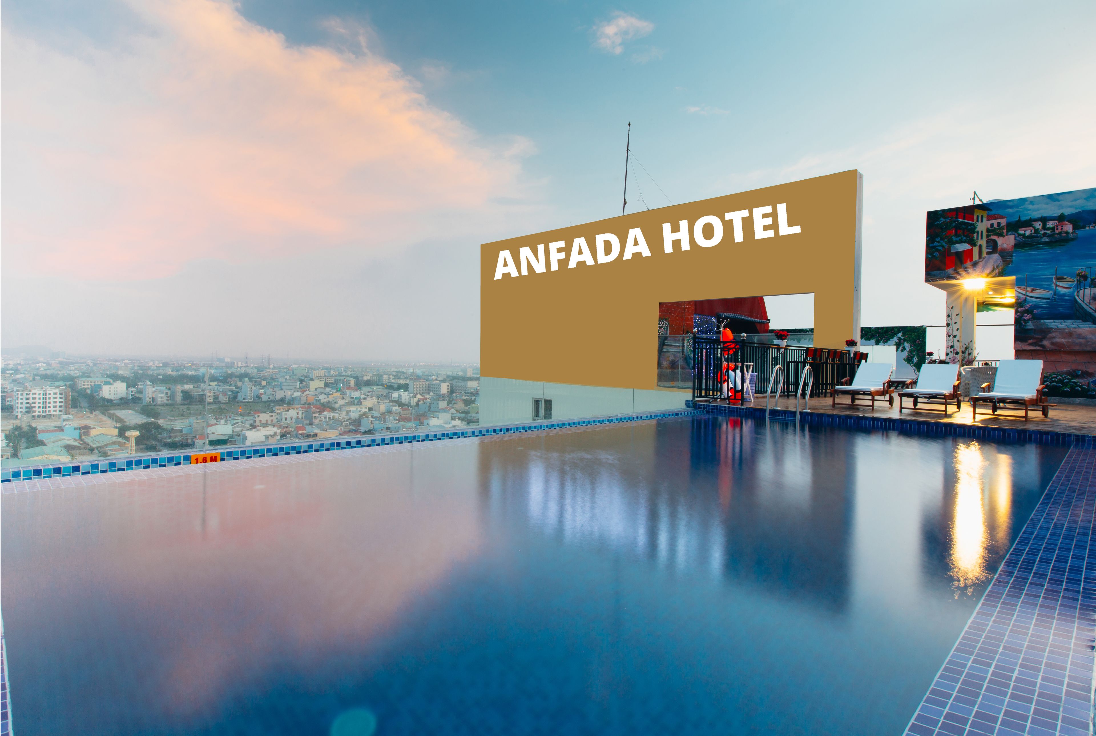 Zona de piscina de hotel con vista a la ciudad, un cartel grande que dice "ANFADA HOTEL" y sillones de descanso.