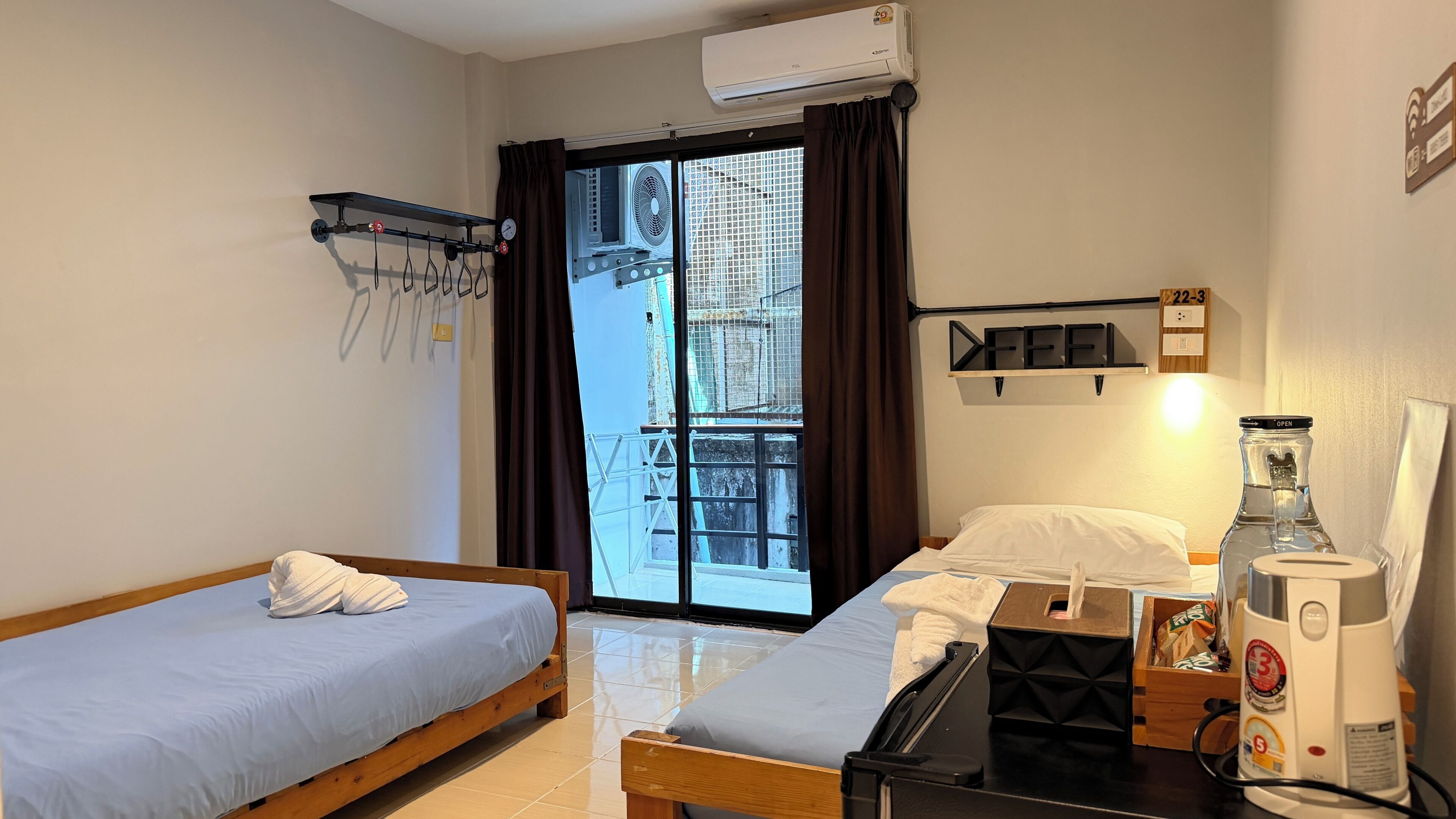A hotel room with two beds, a window with a view of buildings, an air conditioning unit, a shelf with a 'FEEL' sign, and a water dispenser.