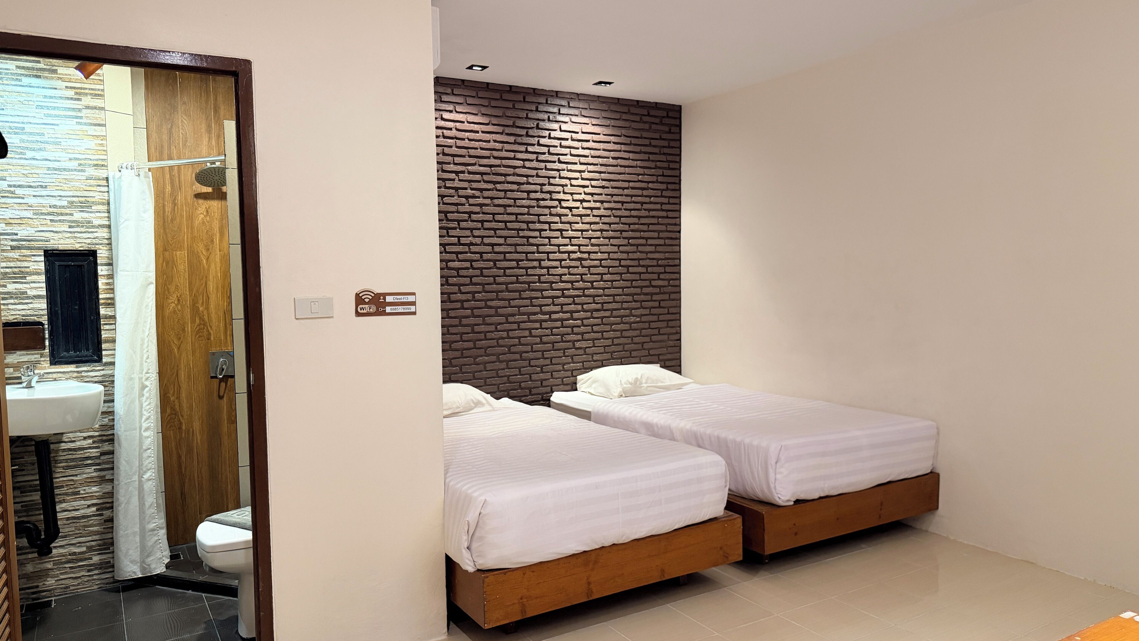 A hotel room with two beds, a wooden headboard, a bathroom with a stone wall, and a visible Wi-Fi sign.
