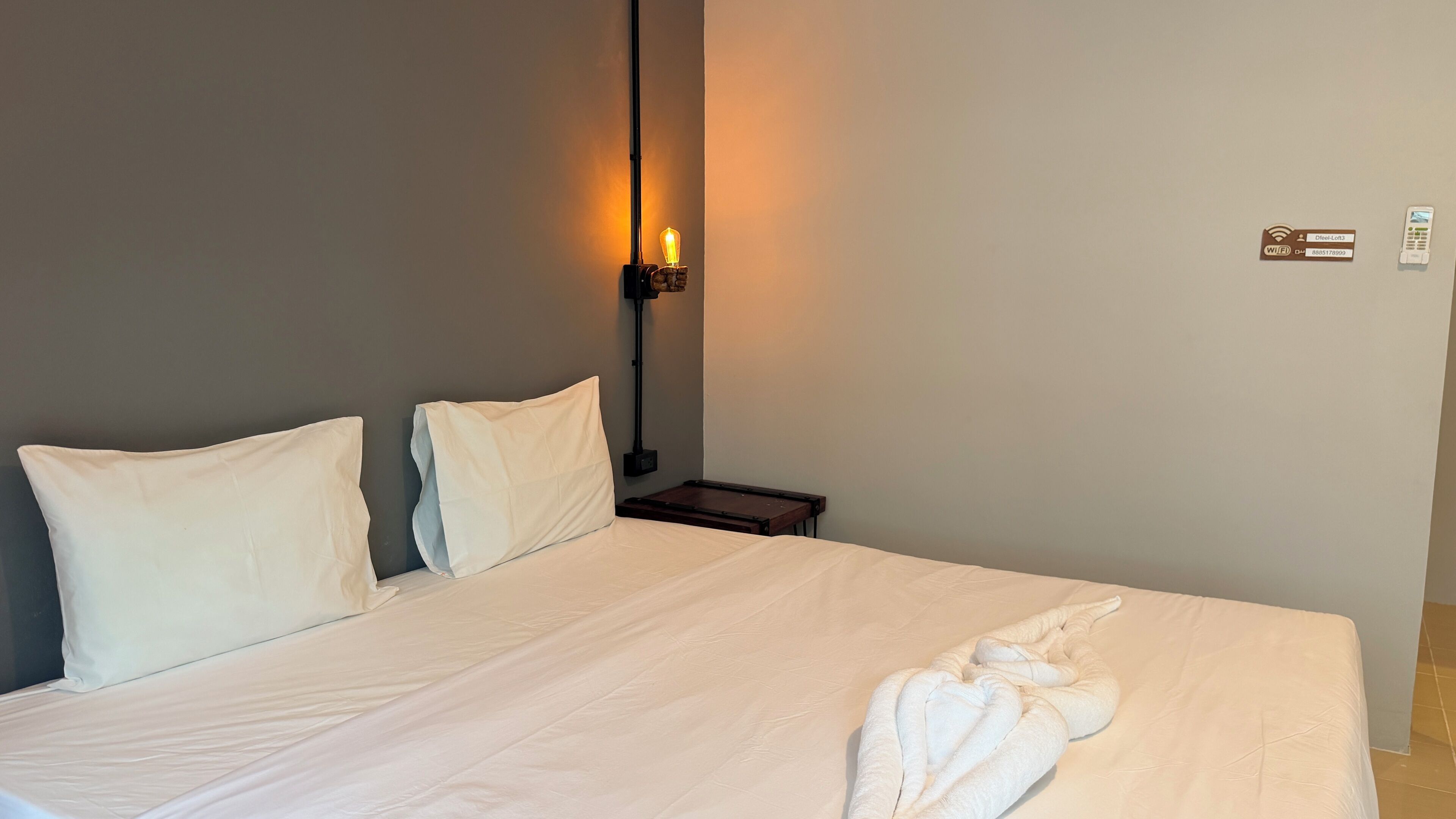 A hotel room with a bed, pillows, a bedside table, a lamp, and a wall-mounted Wi-Fi sign.