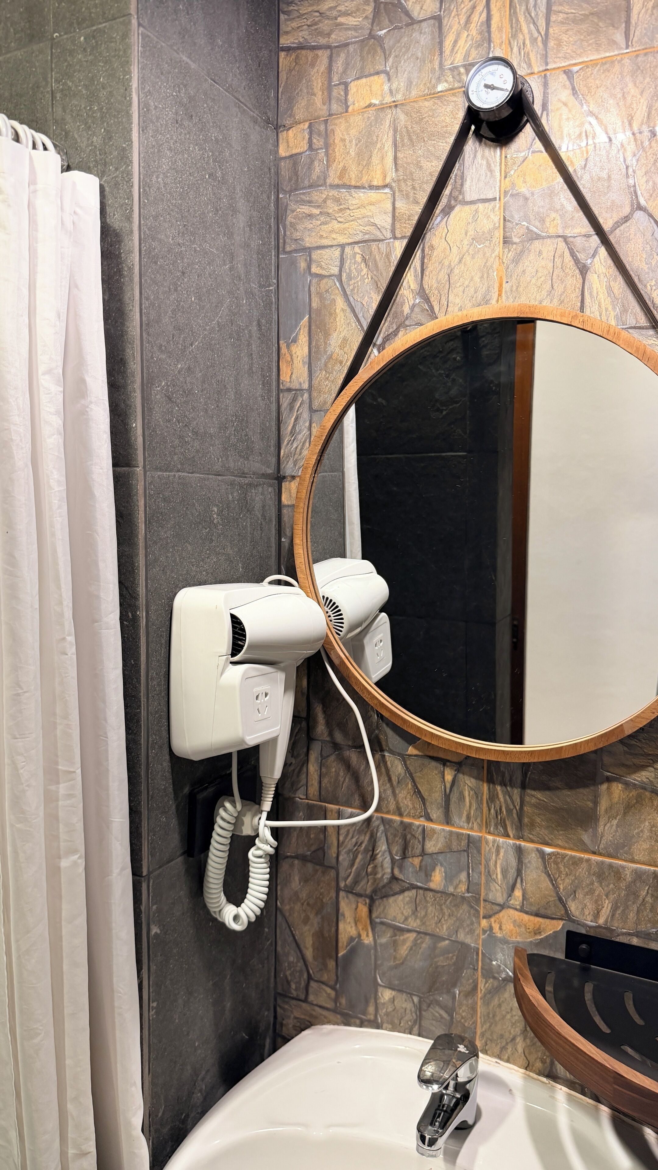 Un bany amb una paret de pedra, un lavabo blanc, un mirall amb estructura de fusta i un assecador de cabells en una estació d'emmagatzematge penjada a la paret.