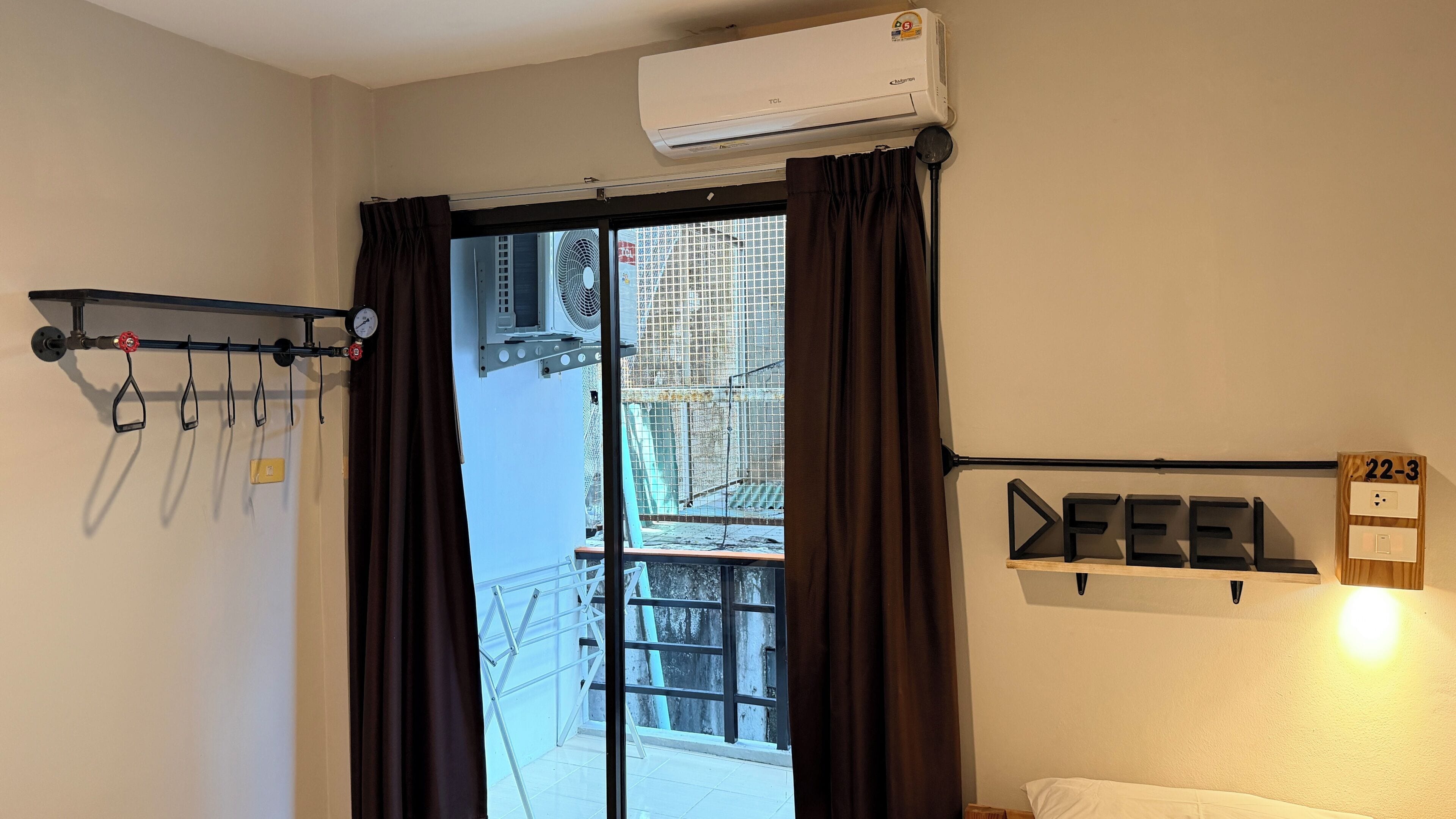 A hotel room with a bed, an air conditioner, a window with curtains, a wall-mounted shelf with letters, and a wall-mounted clock.
