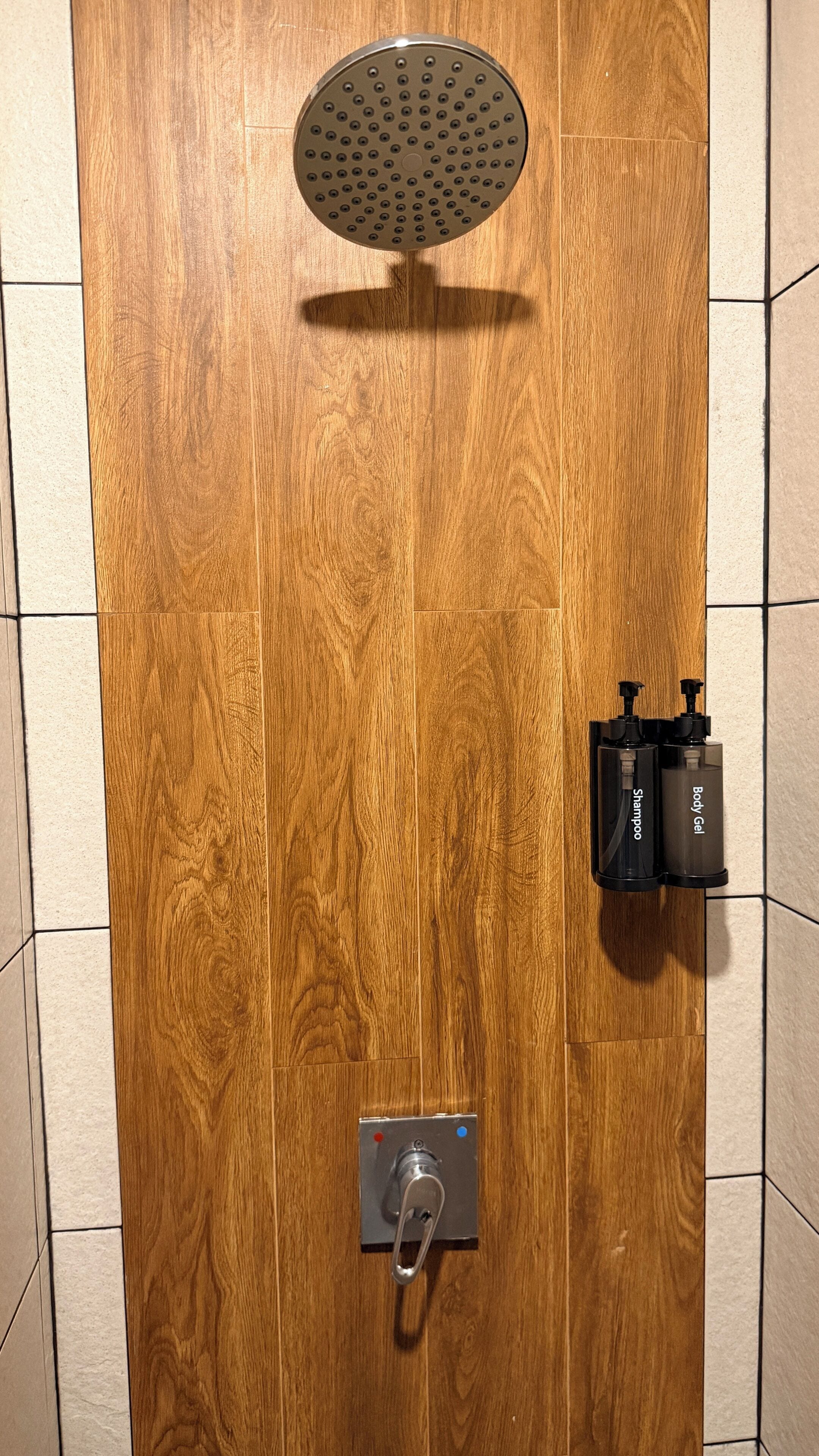 A wooden shower enclosure with a handheld showerhead, a control panel, and two bottles of shampoo and body gel.