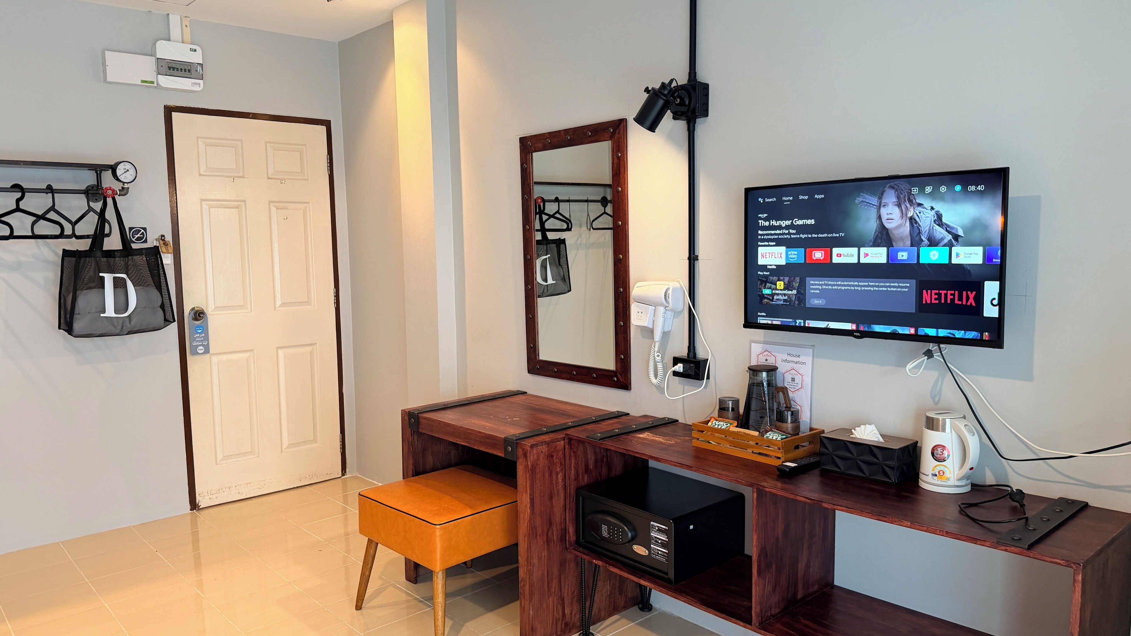 A hotel room with a wooden desk, a television displaying a streaming service, a mirror, and a door with a pressure gauge.