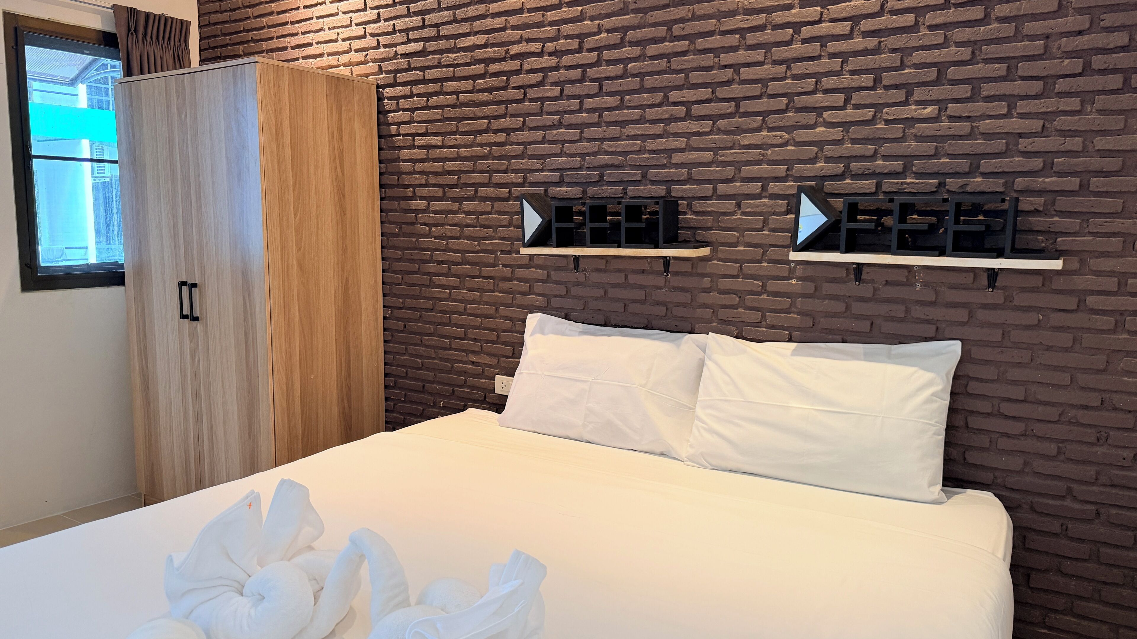 A bedroom with a brick wall, a bed with white bedding, a wardrobe, and a window.