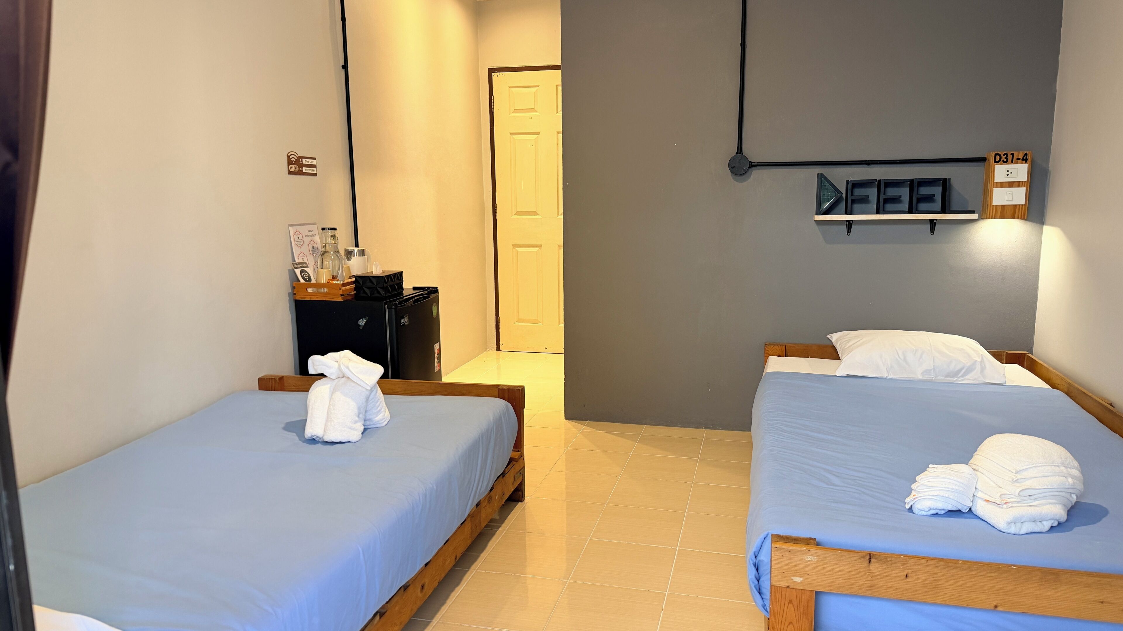 Habitación de hotel con dos camas, un escritorio pequeño con refrigerador, y una puerta que da a un pasillo.