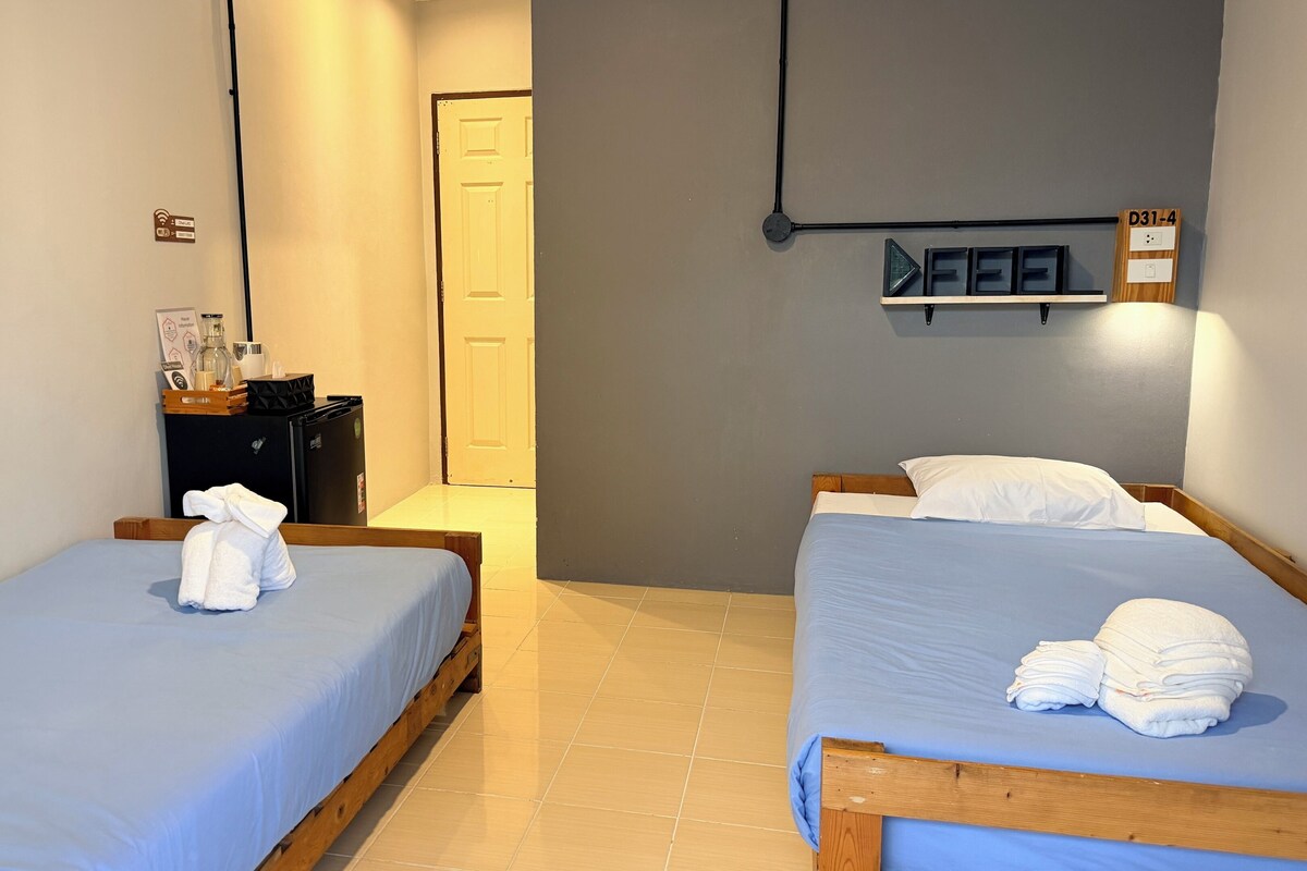 A hotel room with two beds, a small desk with a refrigerator, and a door leading to a hallway.