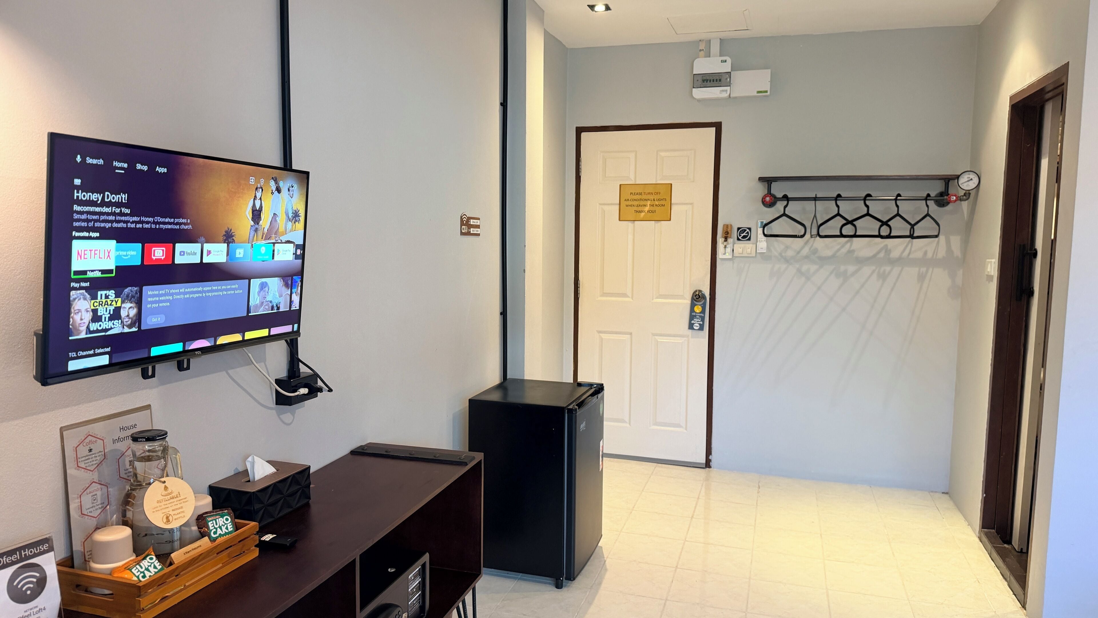 A hotel room with a flat-screen TV mounted on the wall, a small black trash bin, a wooden side table with a tissue box and a glass container, and a door with a sign above it.