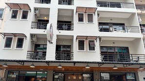 Un immeuble à plusieurs étages doté de balcons, un commerce situé au rez-de-chaussée avec un espace pour s’asseoir, et une vitrine où sont exposés des appareils électroménagers.