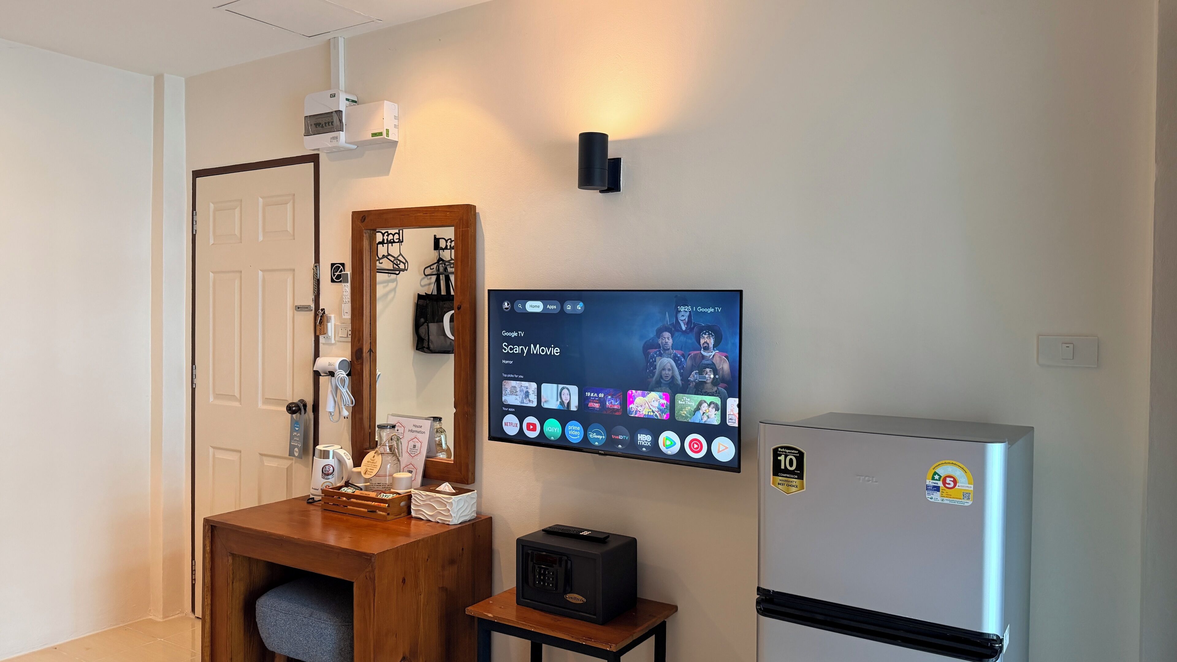 A hotel room with a flat-screen TV displaying a movie selection, a wooden side table with various items, a refrigerator, and a wall-mounted air conditioning unit.