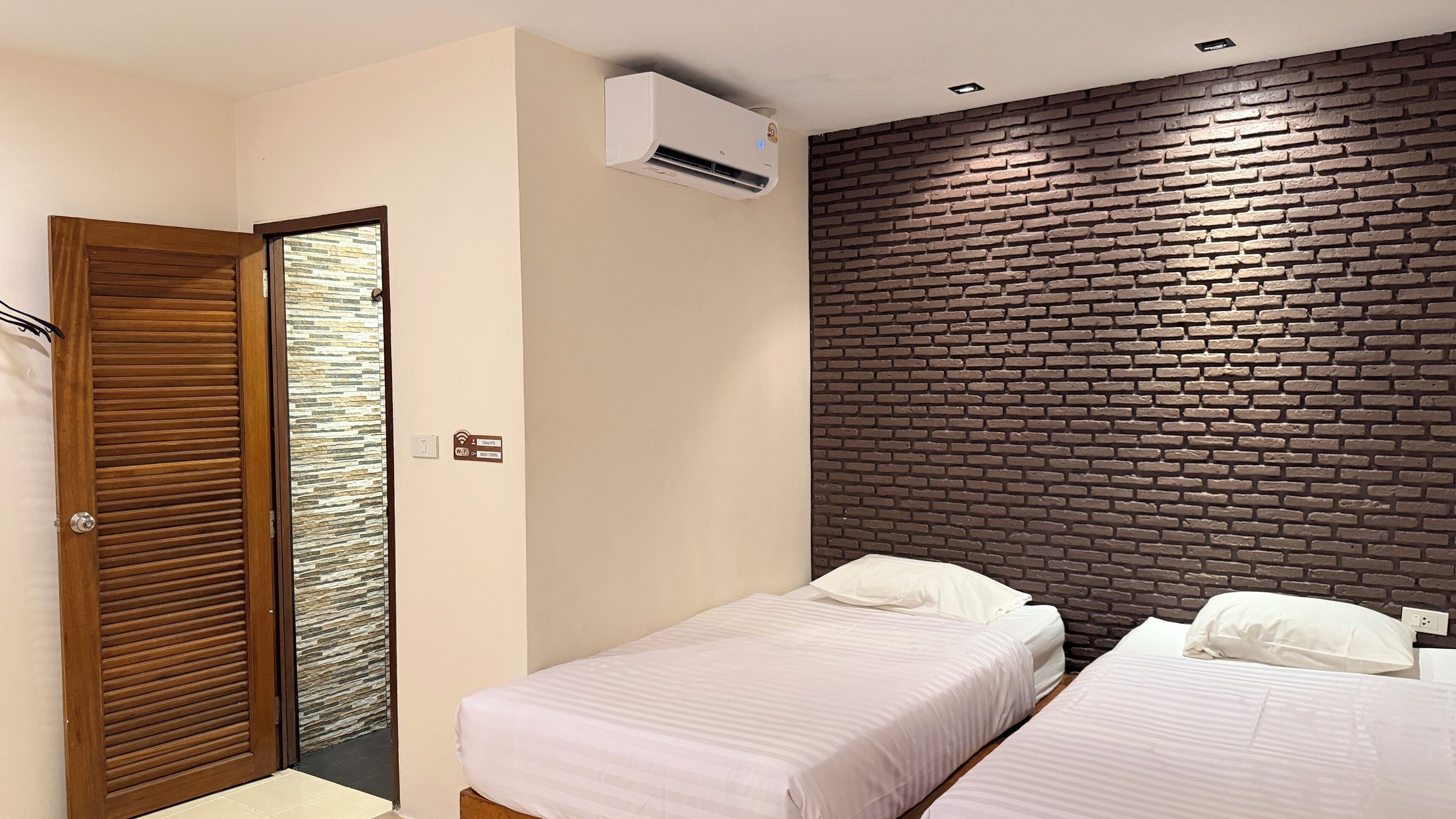 Una habitación de hotel con dos camas, una puerta de madera, un muro con diseño de ladrillo y un equipo de aire acondicionado.