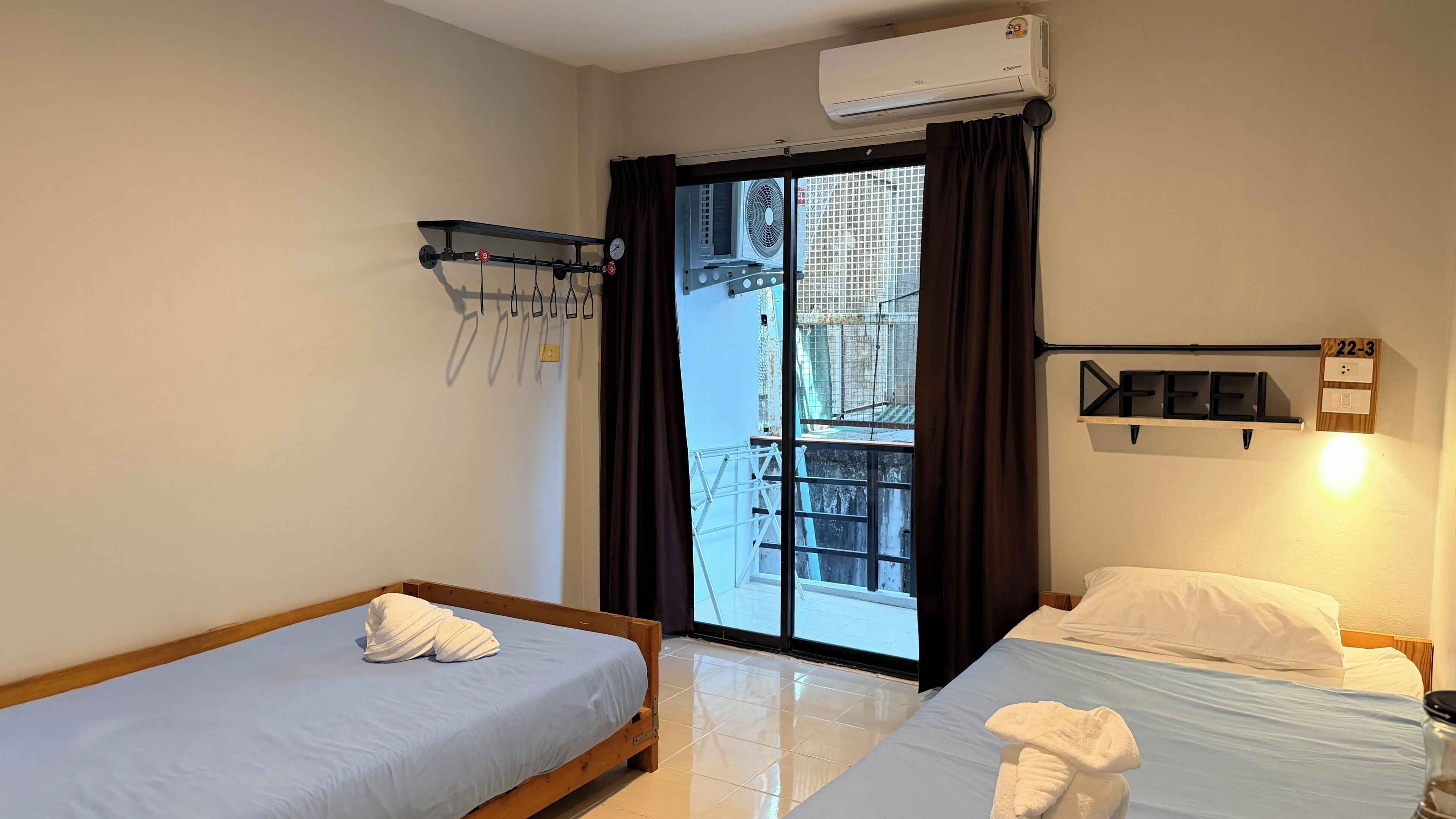 A hotel room with two beds, a window with a view of a building, an air conditioning unit, and a shelf with a towel.
