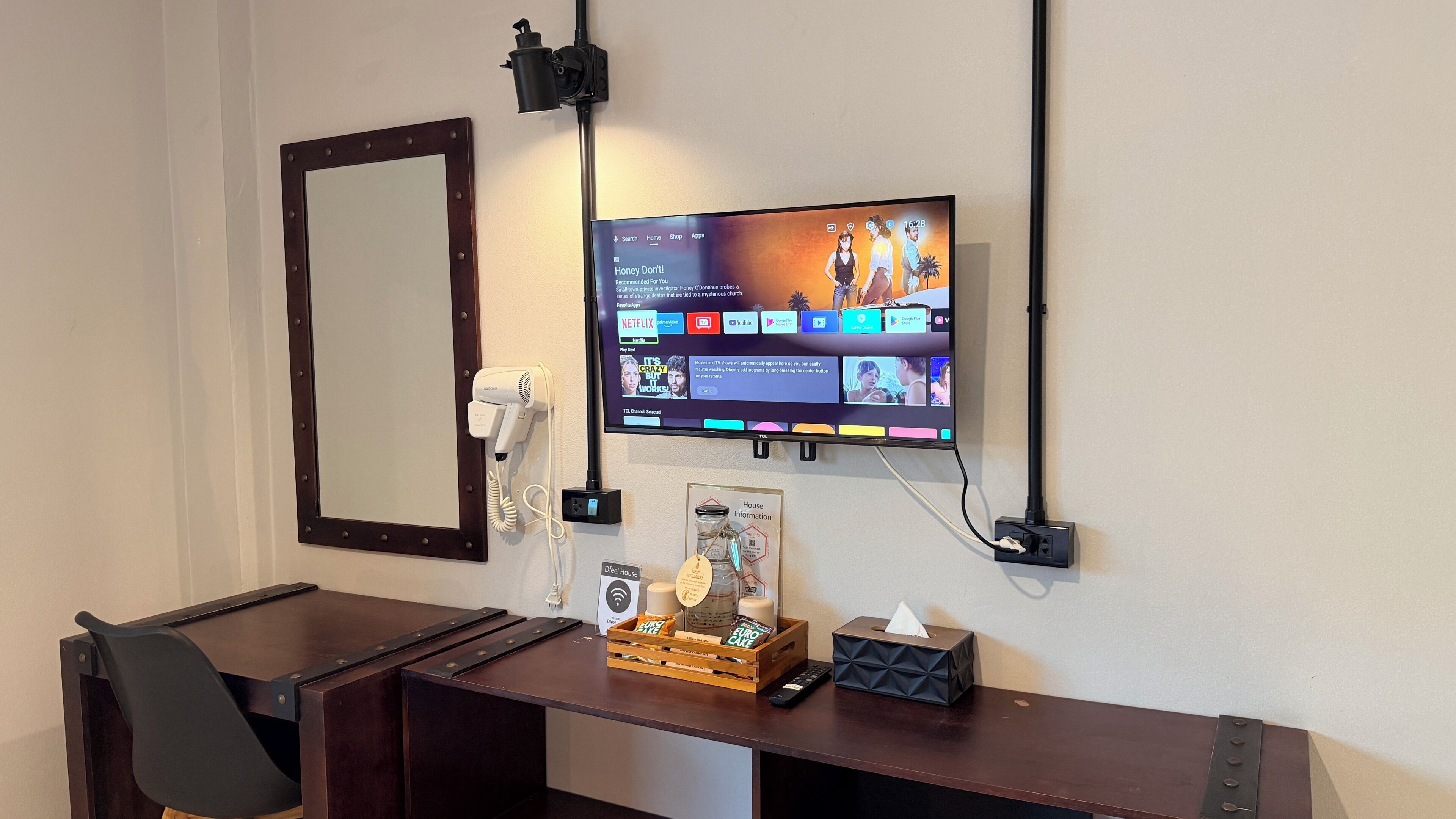 A hotel room with a mounted TV displaying various streaming services, a hairdryer, a mirror, and a desk with a tissue box and remote control.