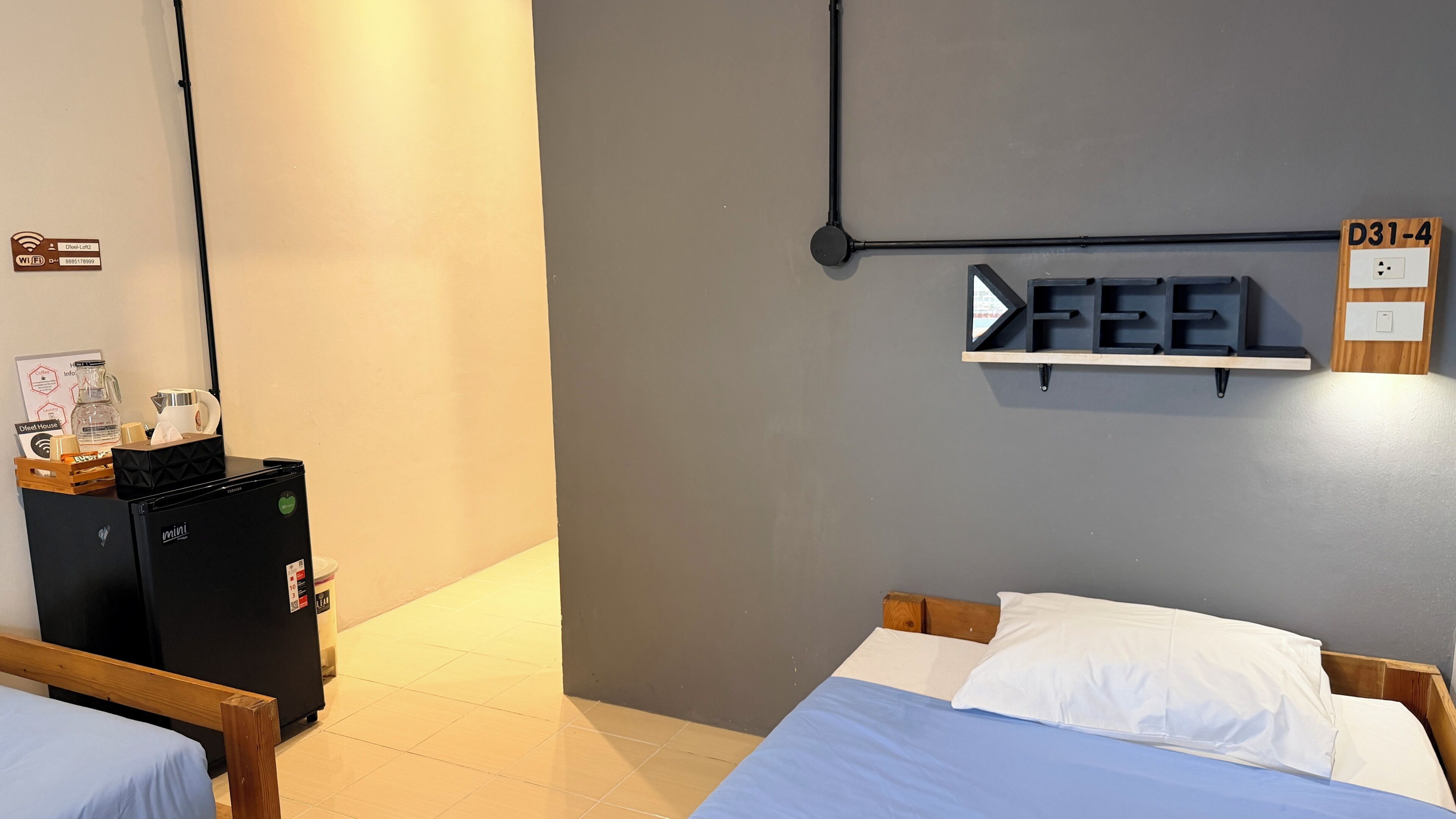 A hotel room with a bed, a desk with a mini-fridge, a coffee machine, and a shelf with a 'FEEL' sign.