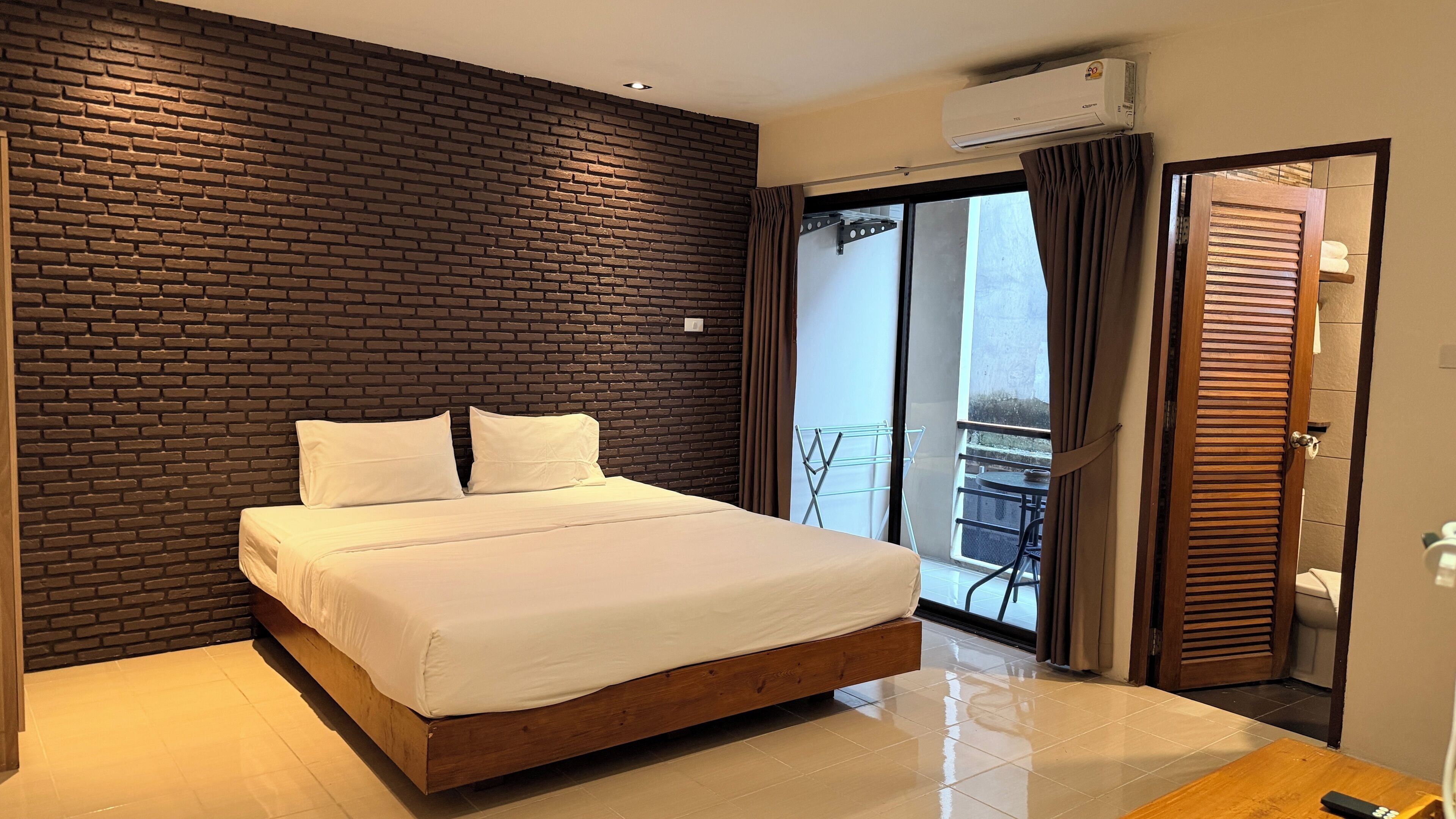 A hotel room with a brick wall, a bed with white linens, a balcony with a chair, and an air conditioning unit.