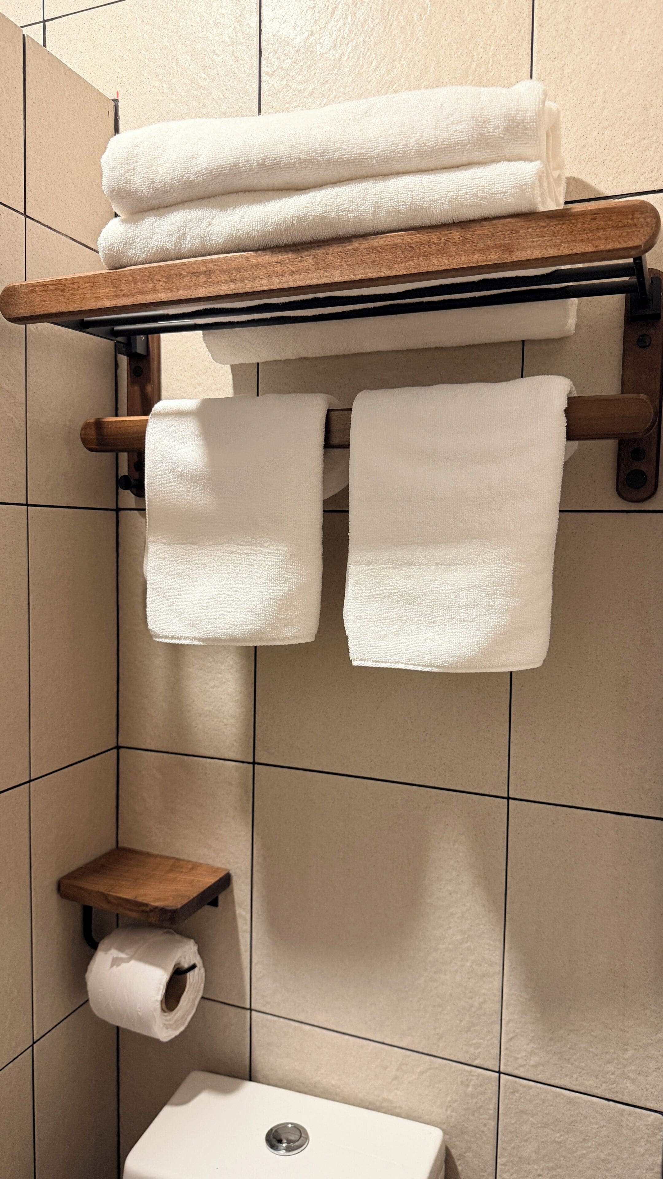 Un baño con un perchero de madera que sostiene tres toallas, un porta papel higiénico y un pequeño estante de madera.