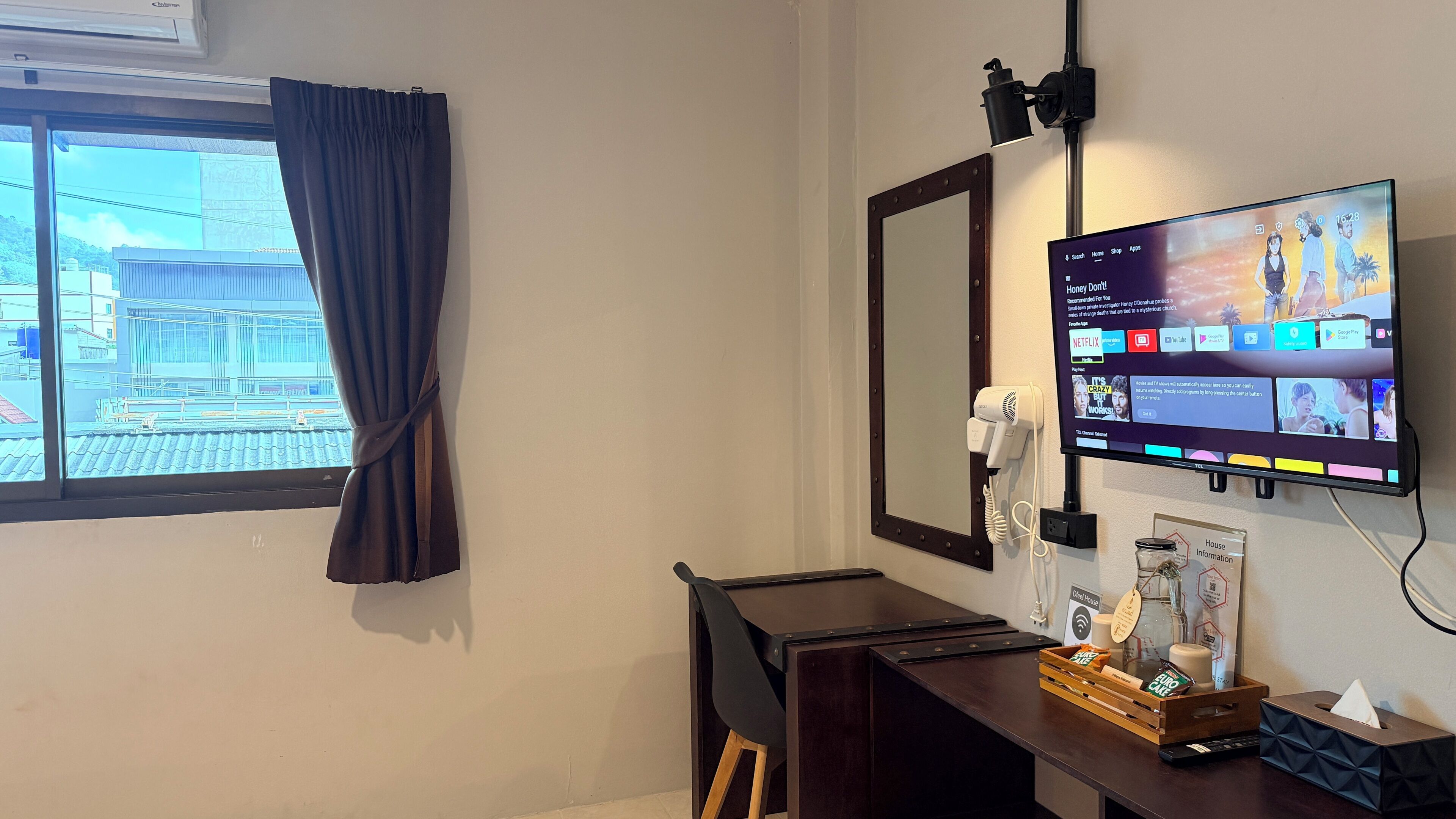 A hotel room with a large window, a flat-screen TV mounted on the wall, a desk with a chair, and a small table with a tissue box and a bottle.