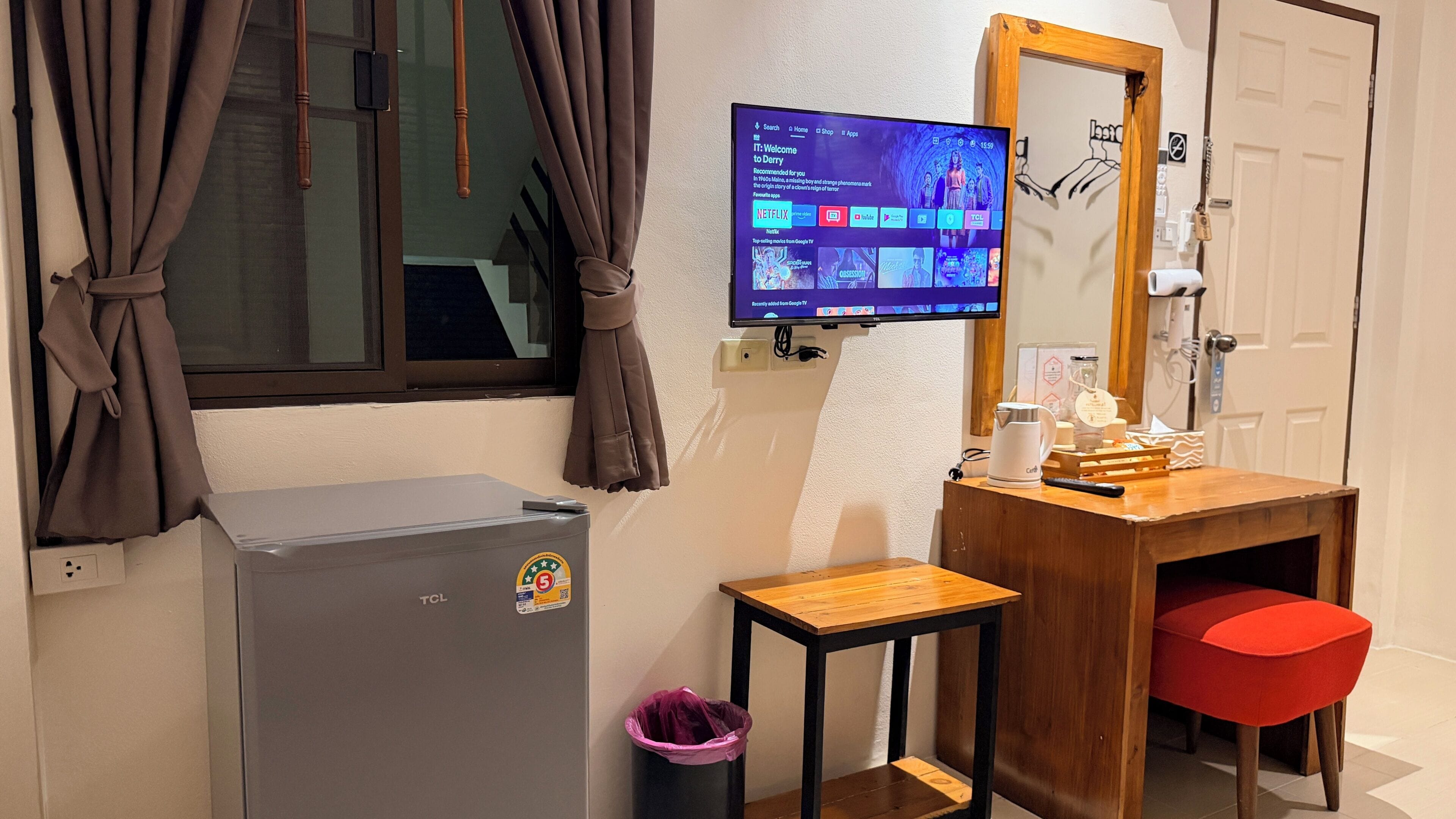 Een kamer met een aan de muur bevestigde televisie waarop een streamingdienst draait, een klein houten tafeltje, een rode stoel, een witte waterkoker en een prullenbak.