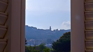 Une vue à travers les volets d’une fenêtre, donnant sur un paysage urbain avec des bâtiments, une tour imposante et un arrière-plan vallonné.