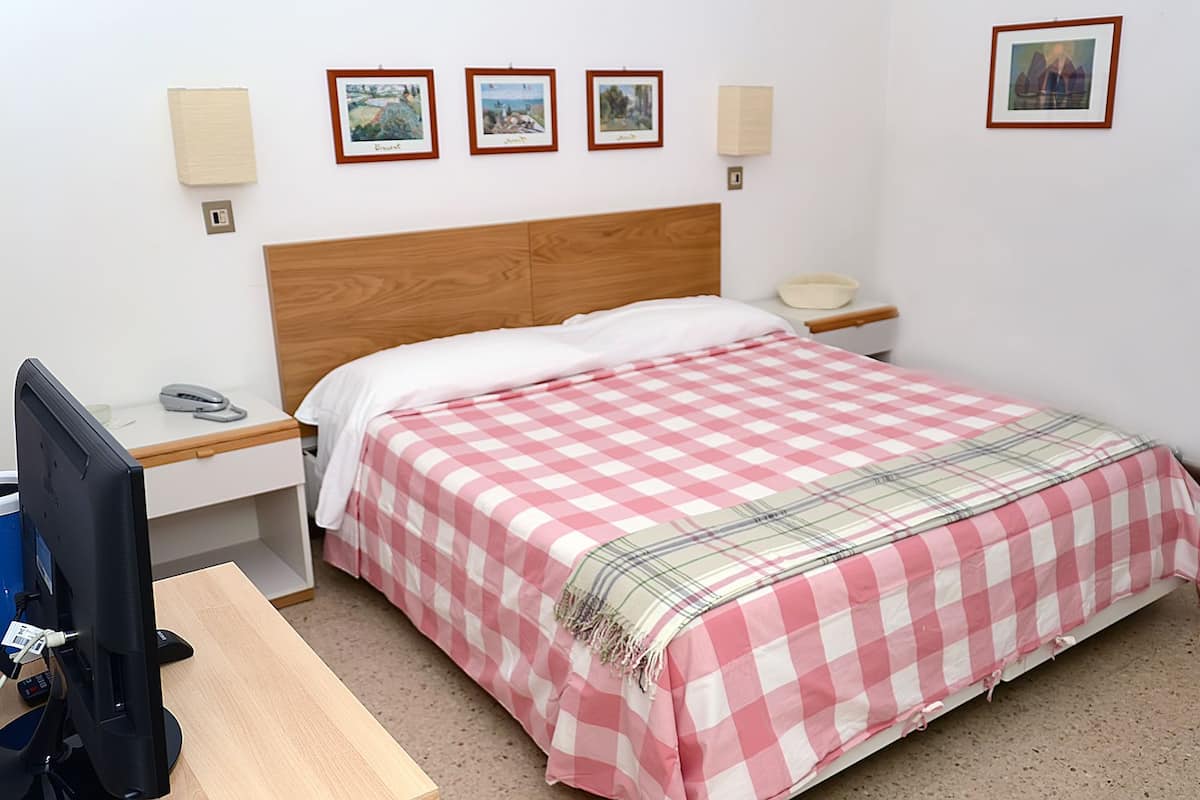 A hotel room with a wooden headboard, a checkered bedspread, a desk with a computer, and a nightstand with a lamp.