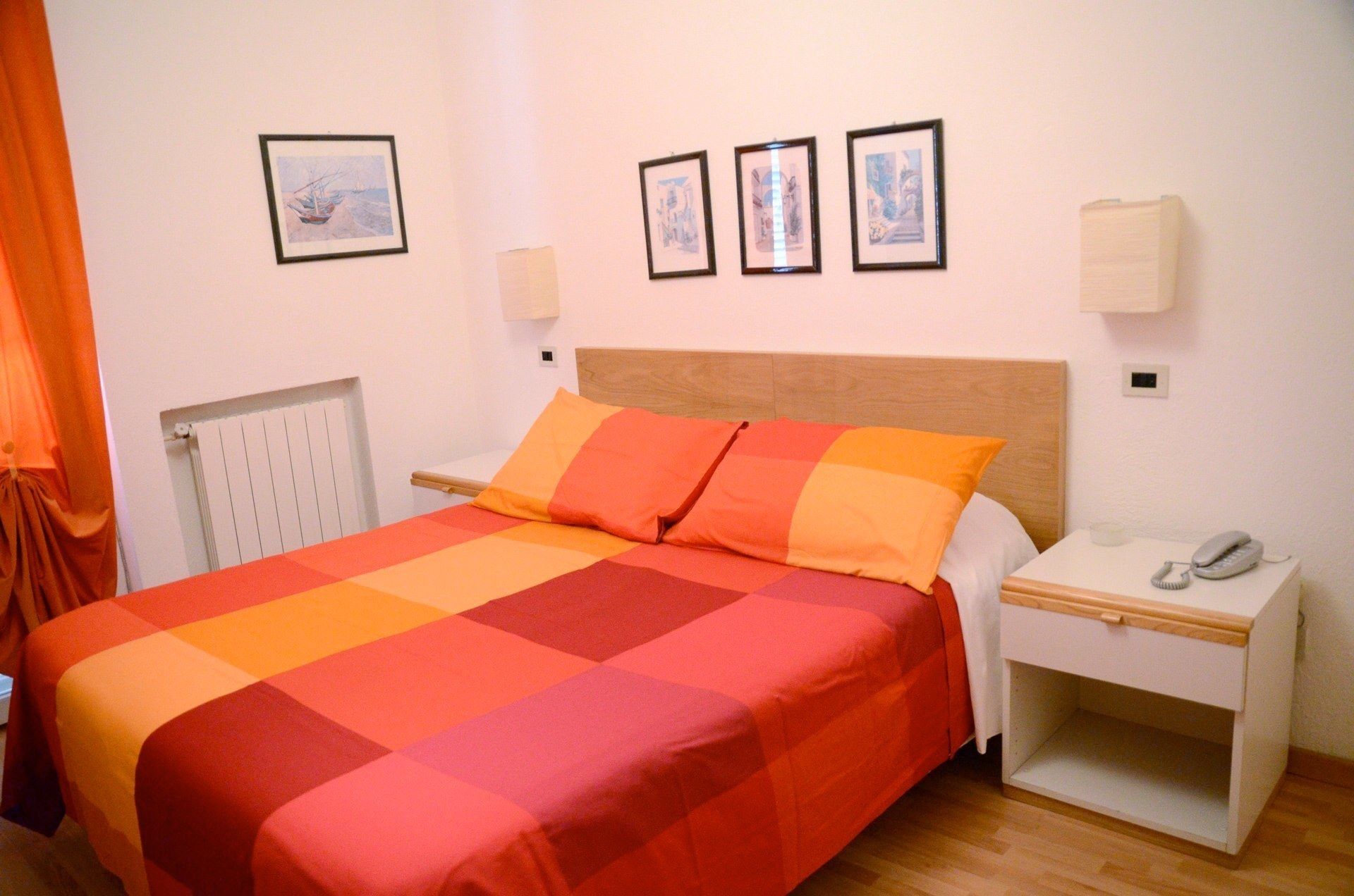 Een netjes opgemaakt bed met een geruit sprei, een nachtkastje met een telefoon, en drie ingelijste foto's aan de muur.