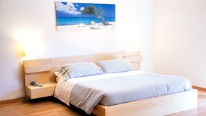 Un lit bien fait, recouvert d’une couvre-lit à rayures, une table de chevet avec une lampe et un tableau représentant une scène de plage accroché au mur.