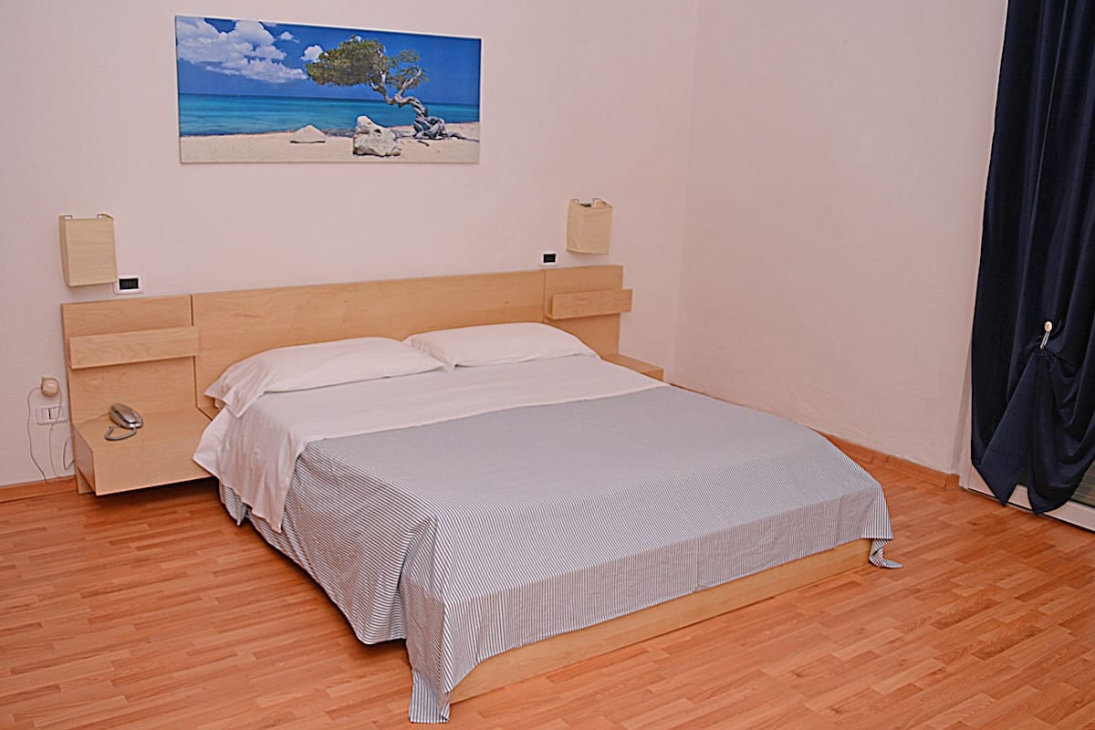 A bedroom with a wooden bed, a nightstand, a bedside table, a painting of a beach scene, and a window with curtains.