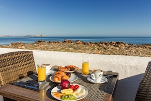 Mesa posta com itens de café da manhã, incluindo croissants, frutas e bebidas, com vista para a costa rochosa.