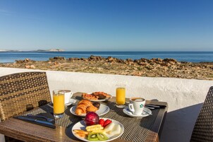 Una mesa dispuesta con productos para el desayuno, como cruasanes, fruta y bebidas, con vista a una costa rocosa.