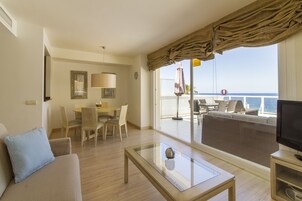 Una sala de estar moderna con sofá, una mesa de centro de vidrio, un comedor con sillas y un balcón con televisor y mobiliario exterior.