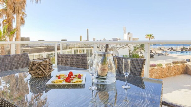 A glass table with a bottle of champagne, a plate of fruit, and two glasses on a balcony overlooking a pool and cityscape. - Can Salia (Sant Josep de sa Talaia)