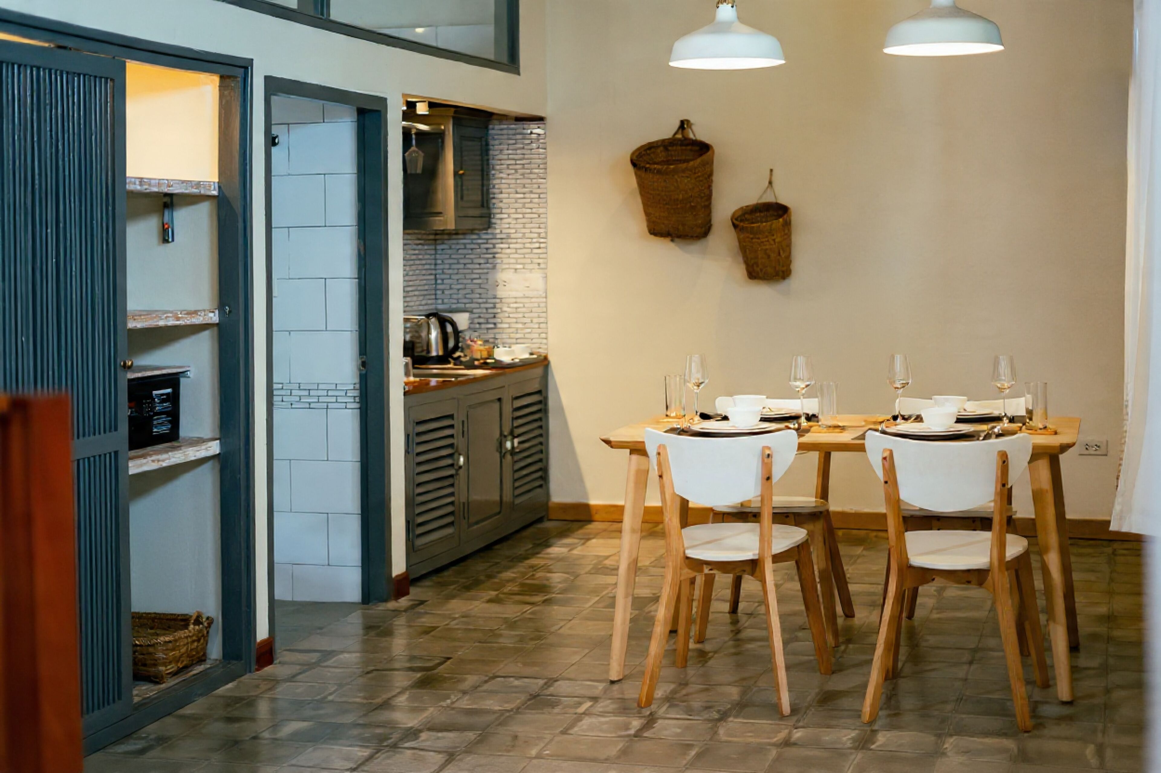 A dining area with a wooden table and chairs, a kitchen in the background, and two hanging baskets on the wall.