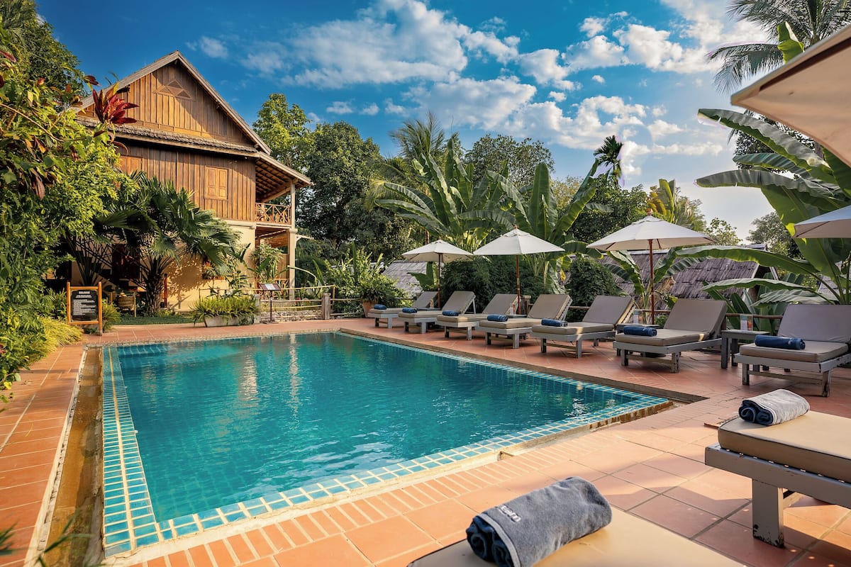Un espace piscine agrémenté de chaises longues et de parasols, entouré de plantes tropicales et d’un bâtiment en bois.