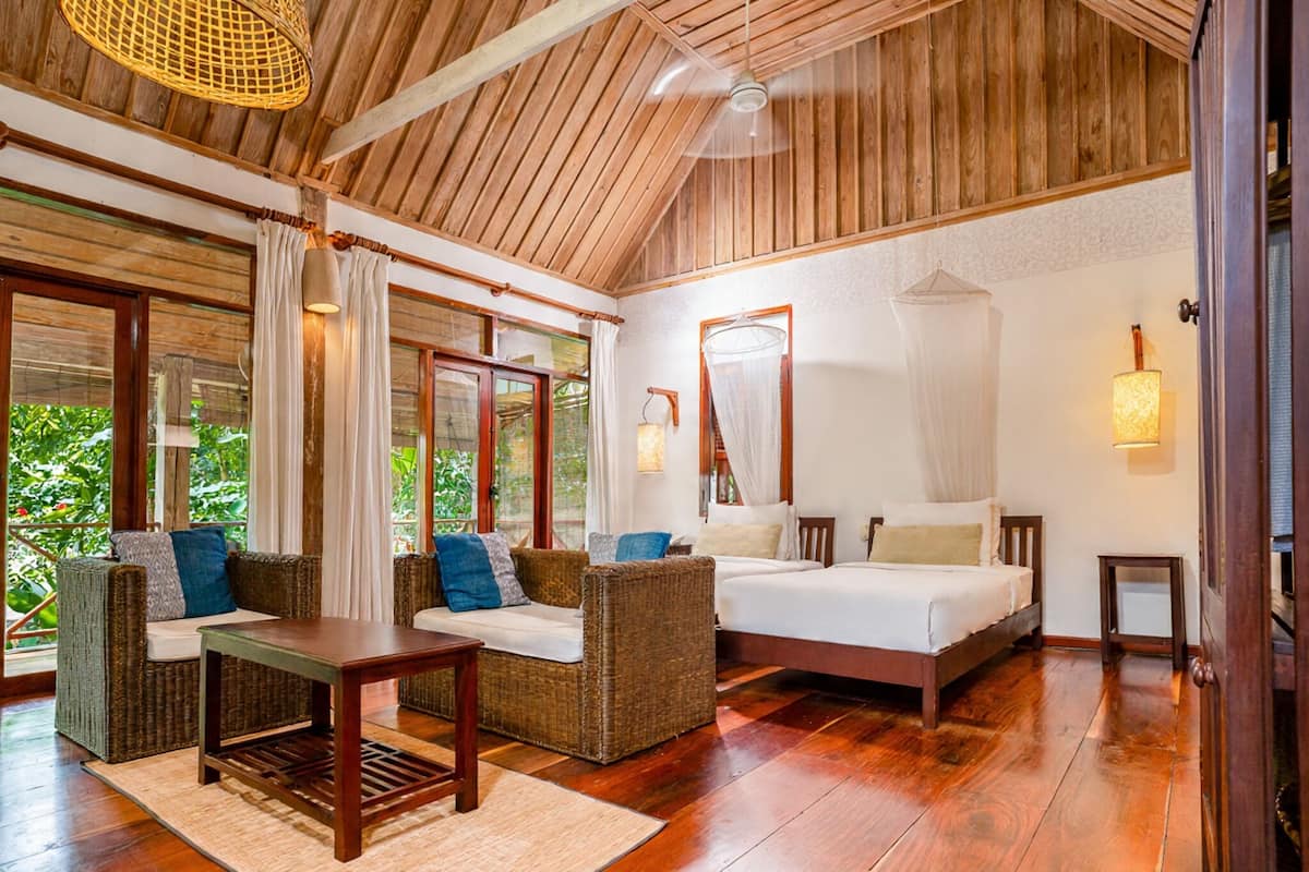 Une pièce spacieuse avec un plafond en bois, un grand lit, deux canapés en osier, une petite table et une vue sur la verdure à l’extérieur.