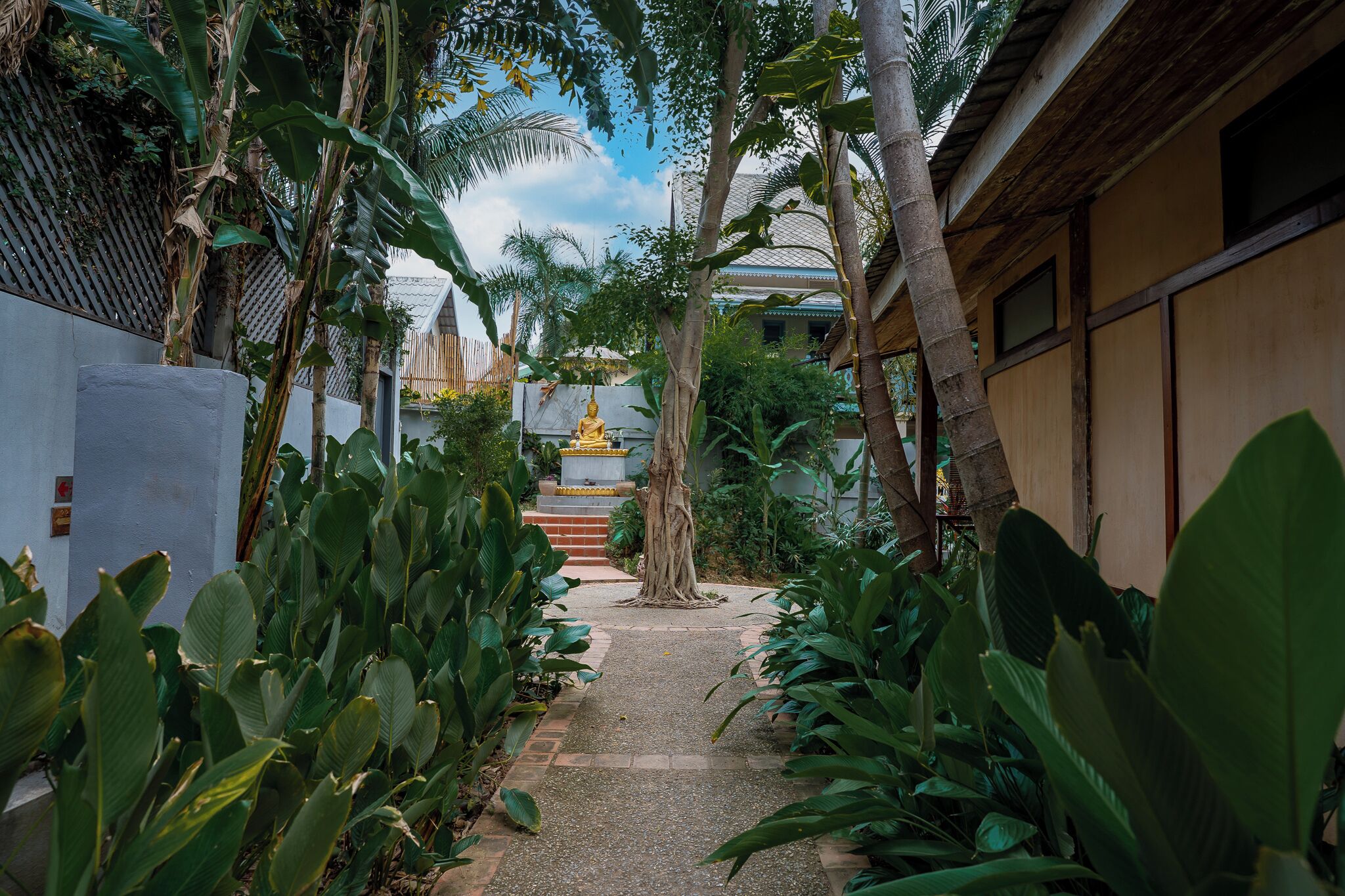 A pathway leading to a golden statue surrounded by lush tropical plants and traditional architecture.