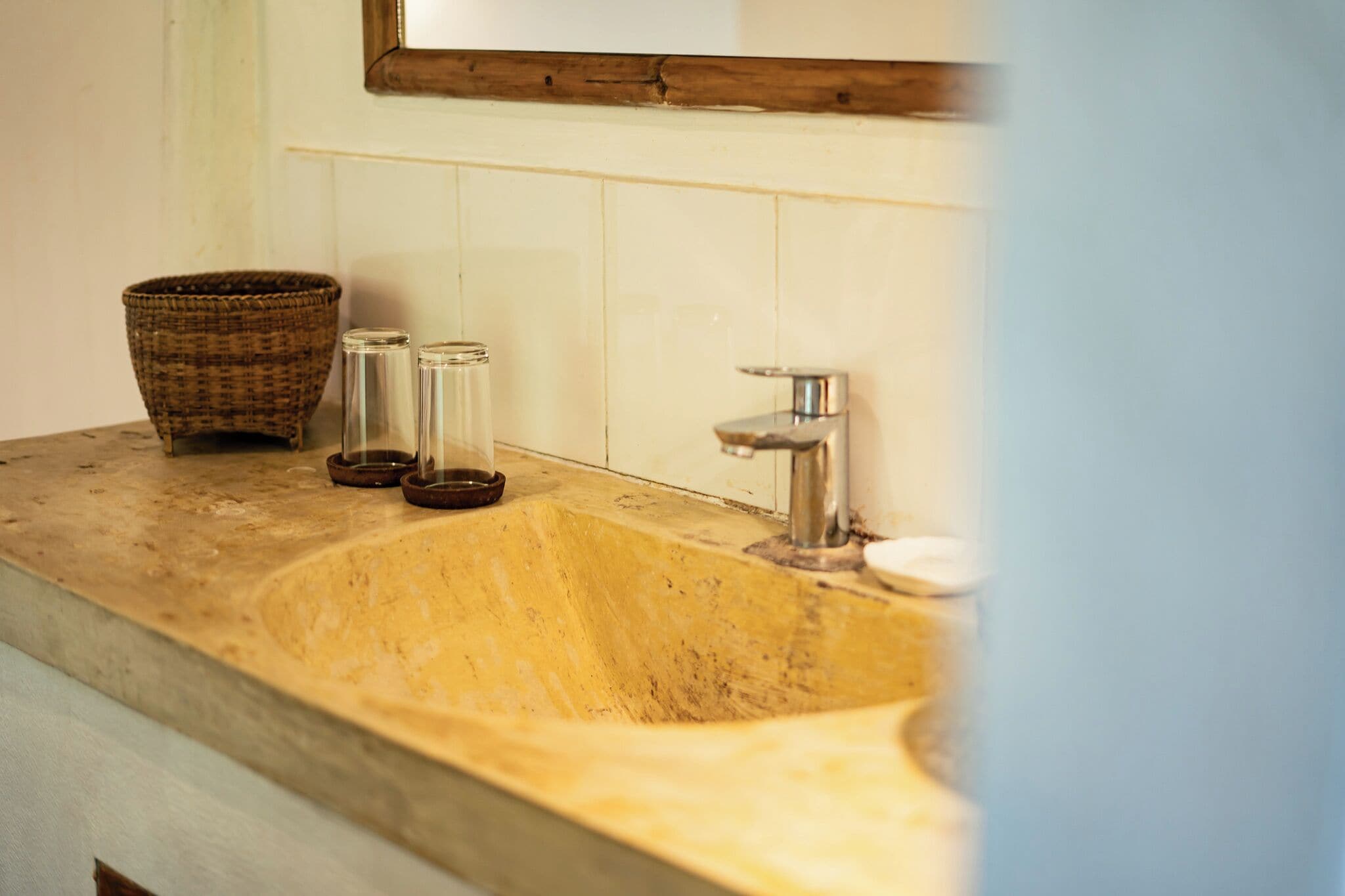 A bathroom sink with a wooden frame mirror, a woven basket, and two glass containers.