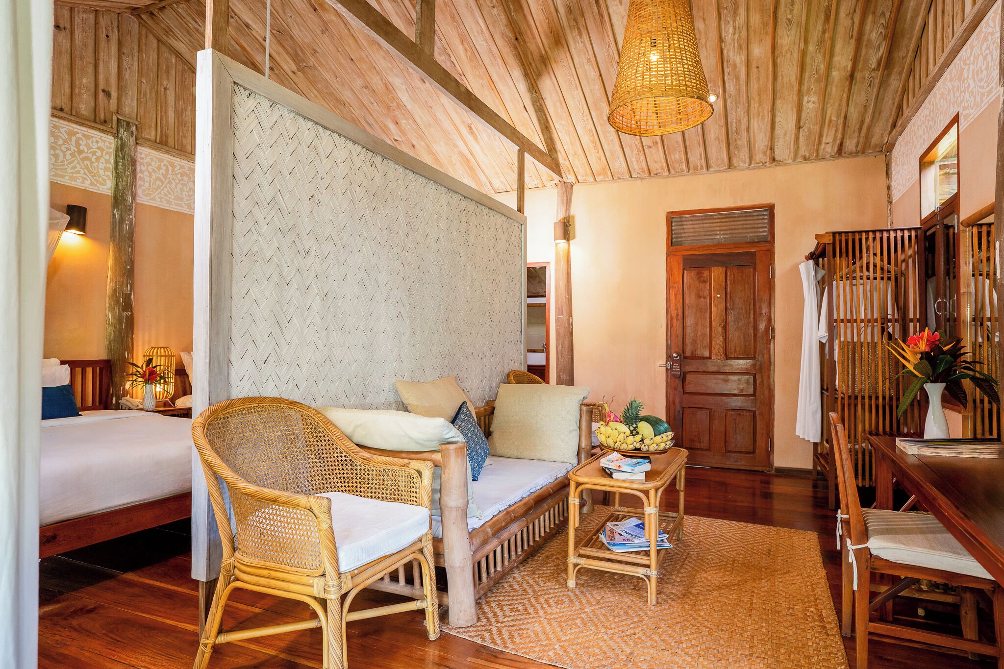 A cozy room with a bed, two wicker chairs, a small table with a fruit bowl, and a woven ceiling light.