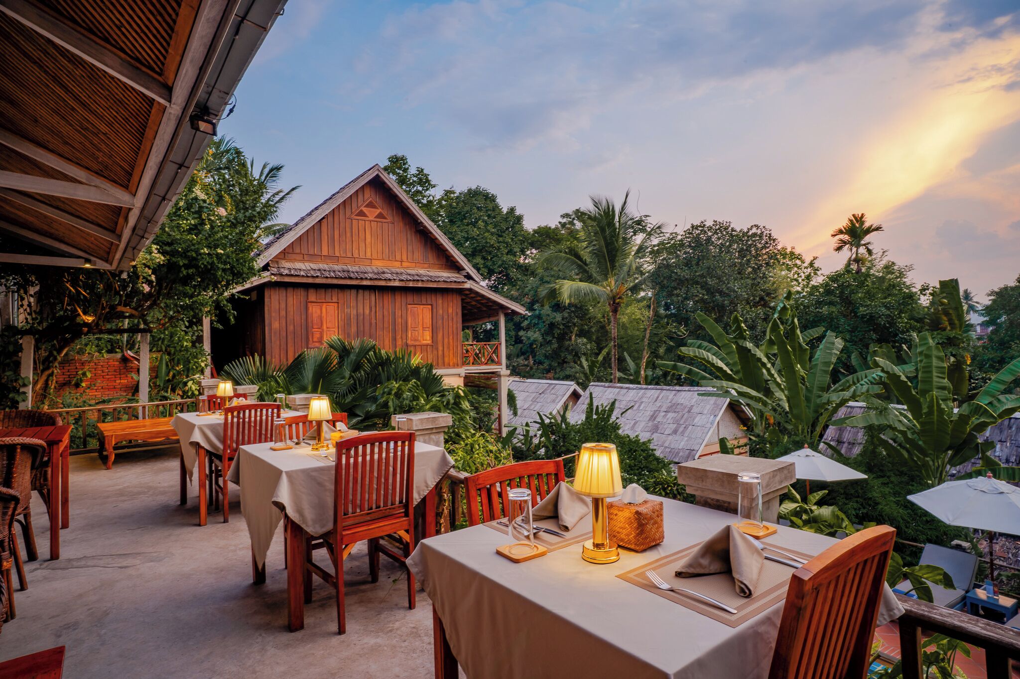An outdoor dining area with wooden tables and chairs, surrounded by lush greenery and a traditional wooden building.