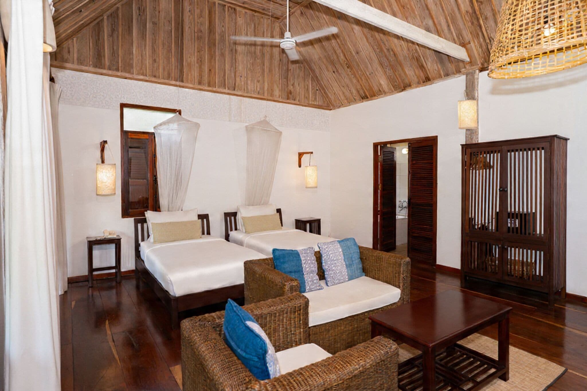 A room with two beds, wicker furniture, a ceiling fan, and a wooden cabinet.
