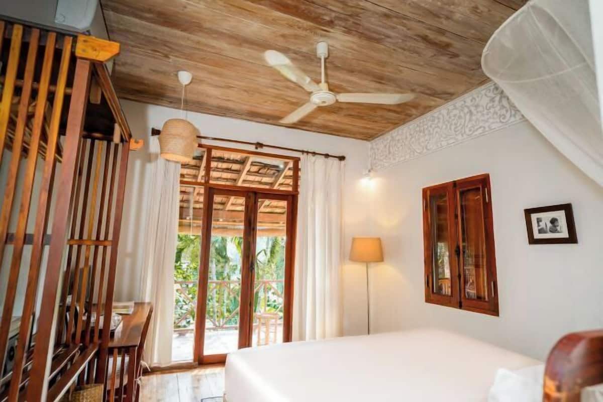 Une chambre à coucher avec un lit, un ventilateur de plafond, une fenêtre avec des rideaux, une lampe de chevet et une chaise en bois.
