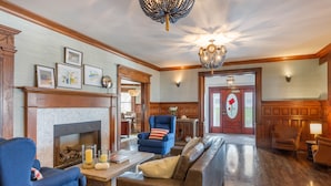 A spacious living room with a fireplace, a blue armchair, a brown sofa, and a chandelier. - The Shore House (Narragansett)