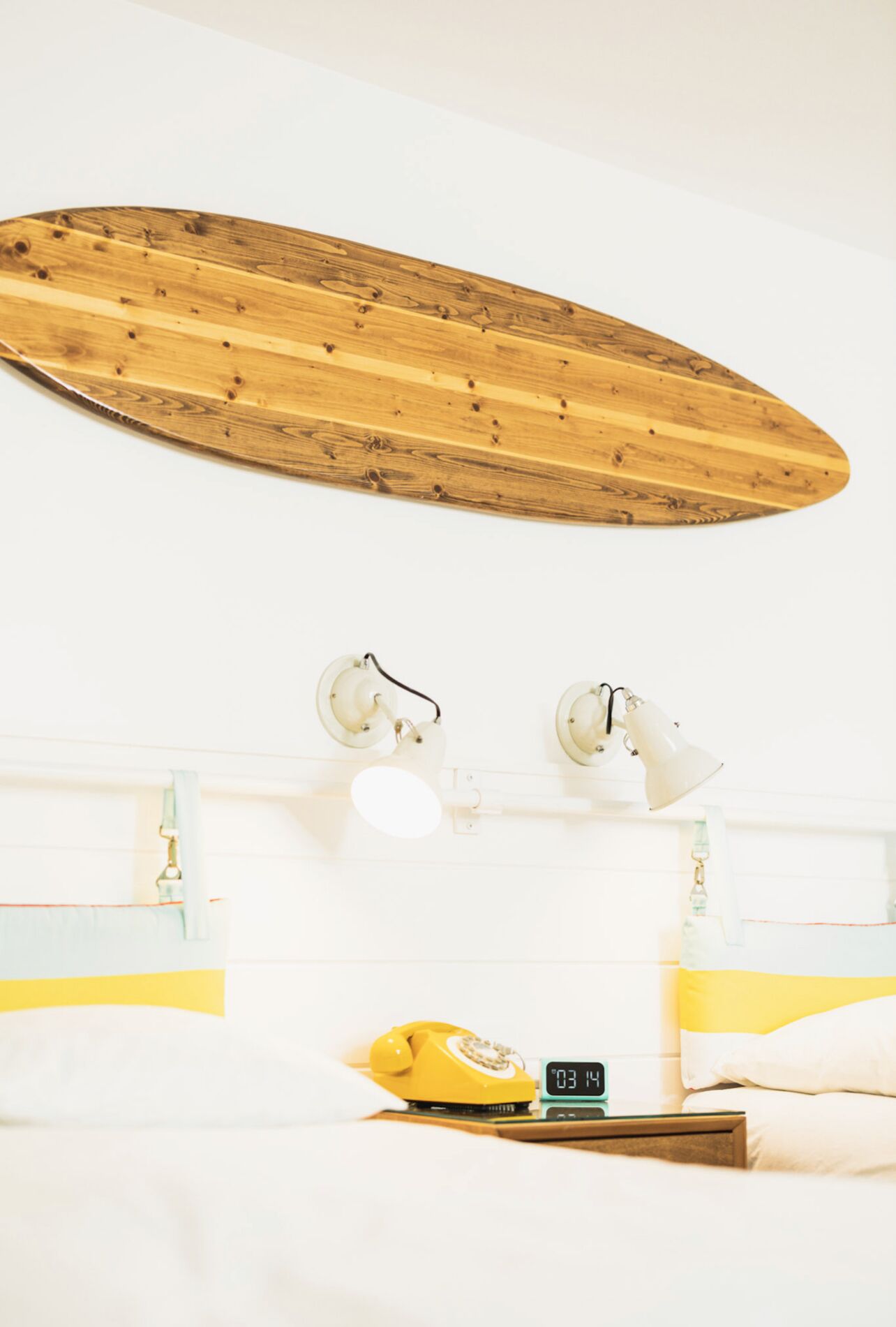 Una habitación con una tabla de surf de madera montada en la pared, una cama con ropa de cama blanca, un teléfono amarillo y un reloj digital que muestra la hora.