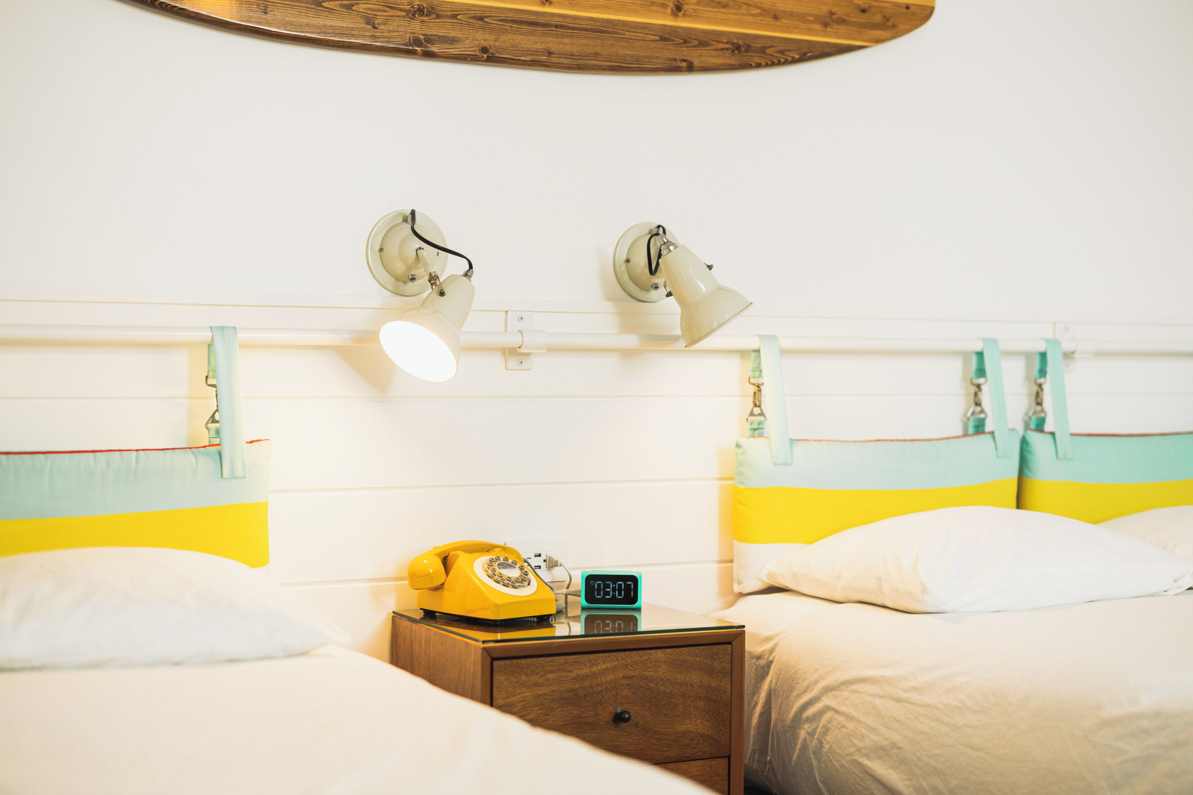 A hotel room with two beds, each with a headboard featuring a striped design. There is a wooden nightstand with a rotary phone and a digital clock.