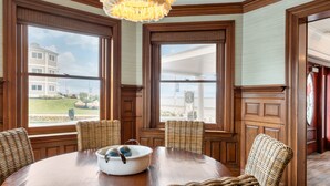 A dining area with a round wooden table, wicker chairs, and a large window with a view of a building and greenery. - The Shore House (Narragansett)