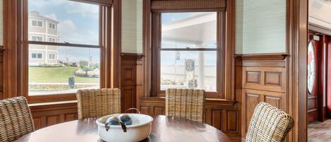 A dining area with a round wooden table, wicker chairs, and a large window with a view of a building and greenery.