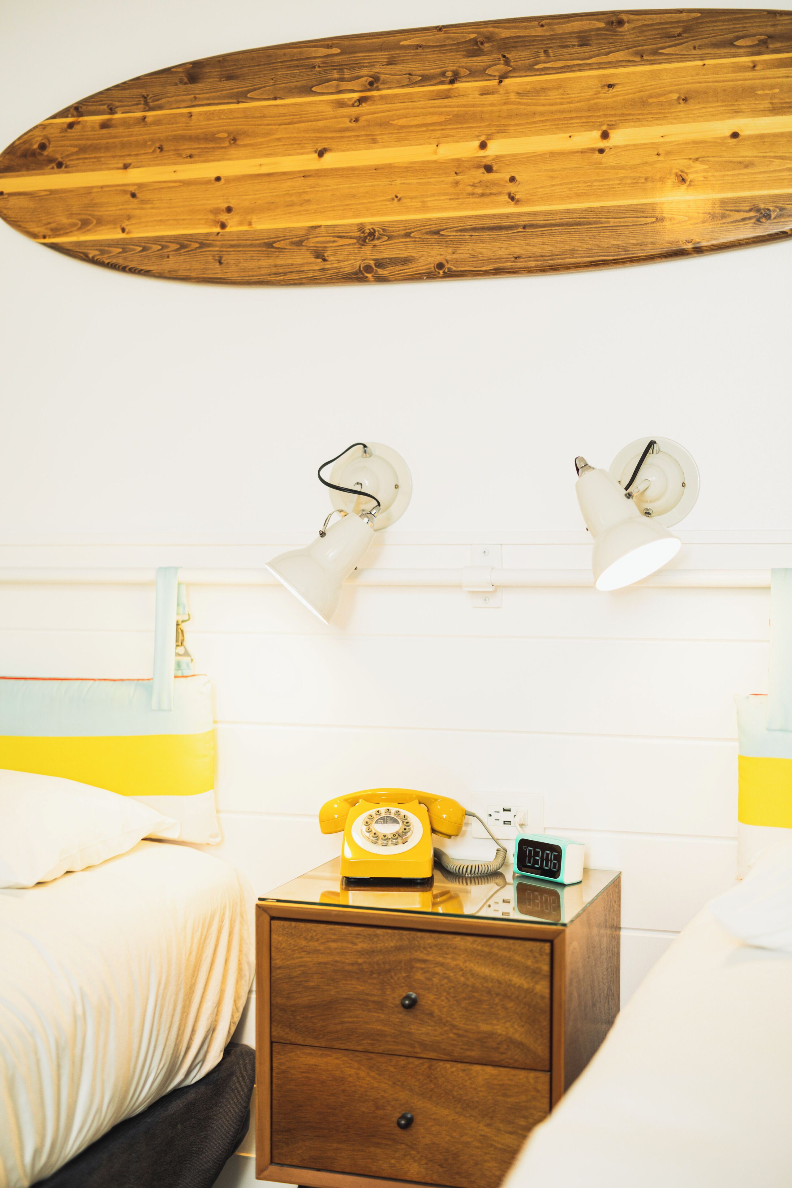 A hotel room with a wooden ceiling, a bed with white bedding, a nightstand with a yellow rotary phone, a digital clock, and two wall-mounted lamps.