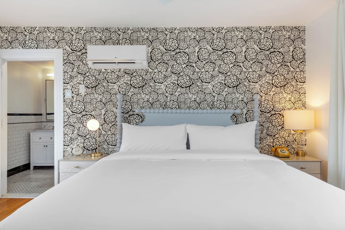 A bedroom with a patterned wallpaper, a bed with white linens, a nightstand with a lamp, and a bedside table with a telephone.
