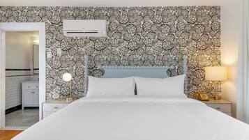 A bedroom with a patterned wallpaper, a bed with white linens, a nightstand with a lamp, and a bedside table with a telephone.