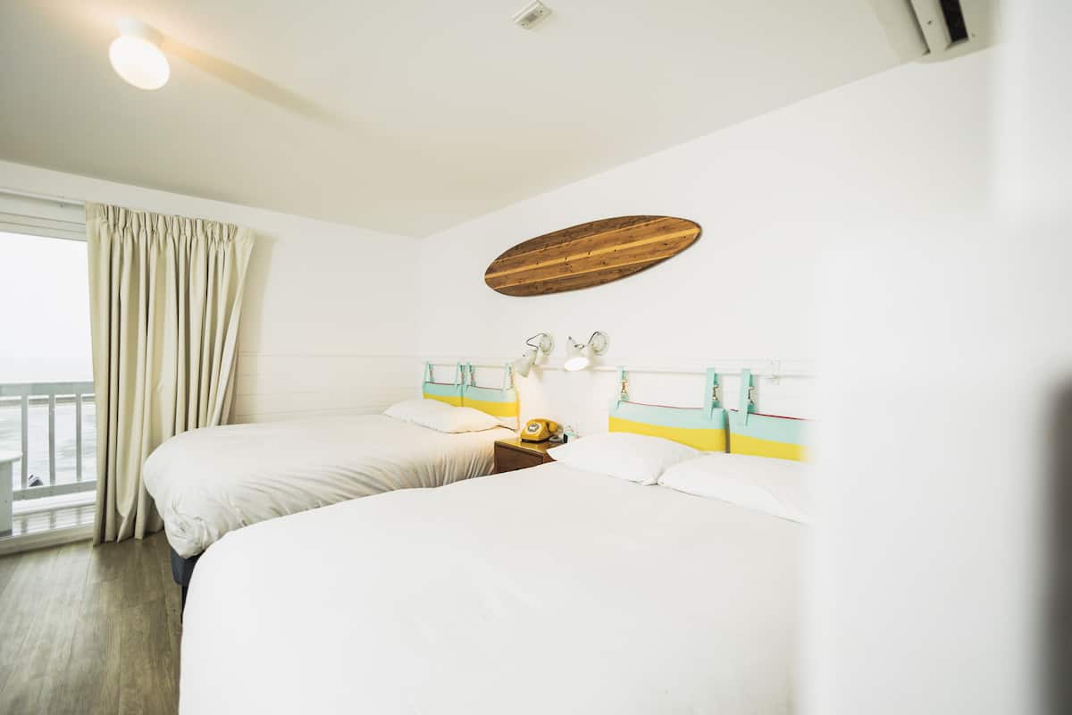 A hotel room with two beds, a wooden headboard, a nightstand with a lamp, and a window with curtains.
