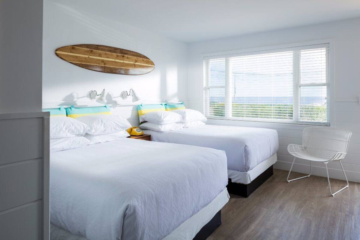 A bedroom with two beds, a wooden surfboard wall art, a white chair, and a large window with blinds.